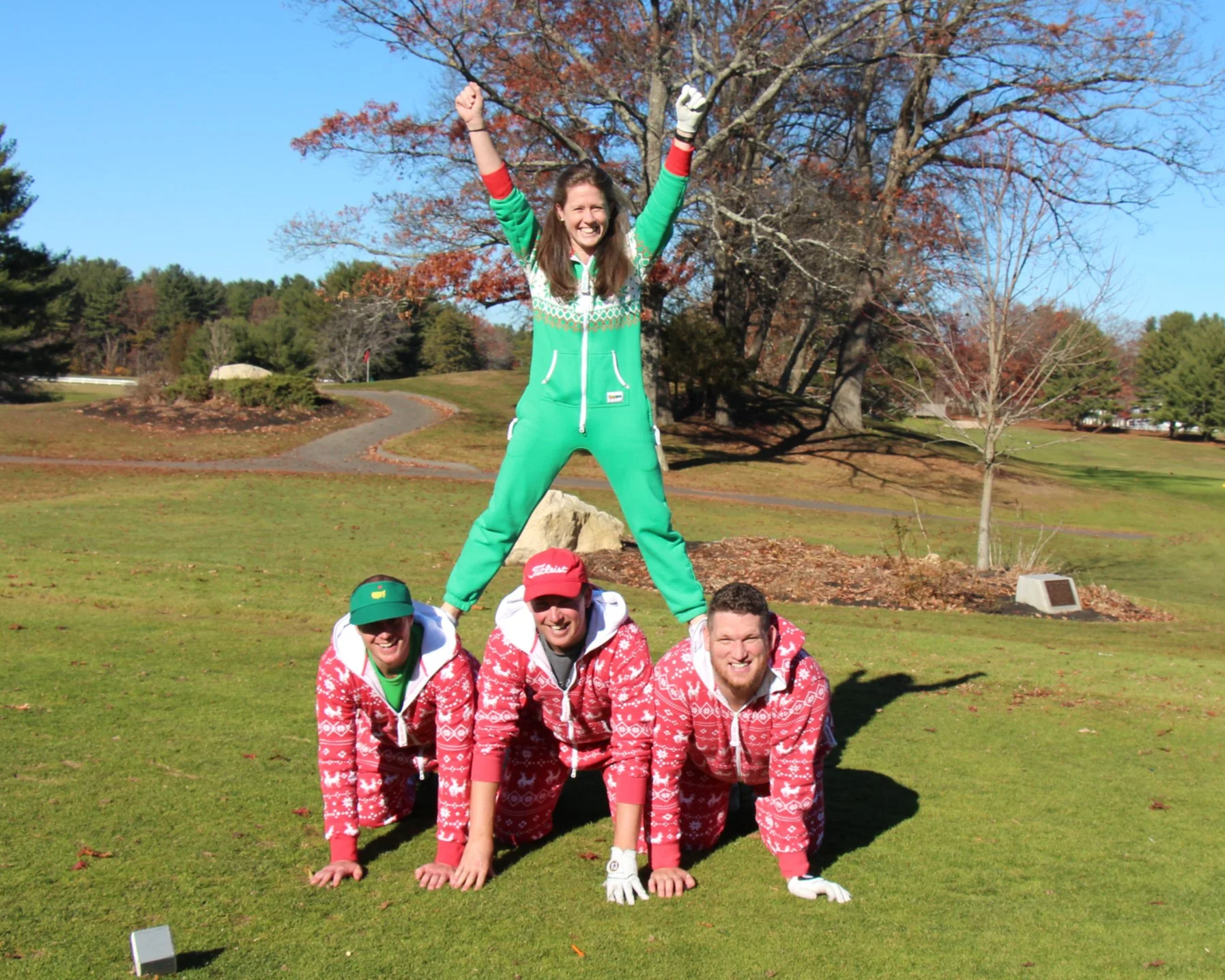 Four people in festive red and green outfits pose outdoors on a sunny grassy lawn, with one person standing on the others’ backs.