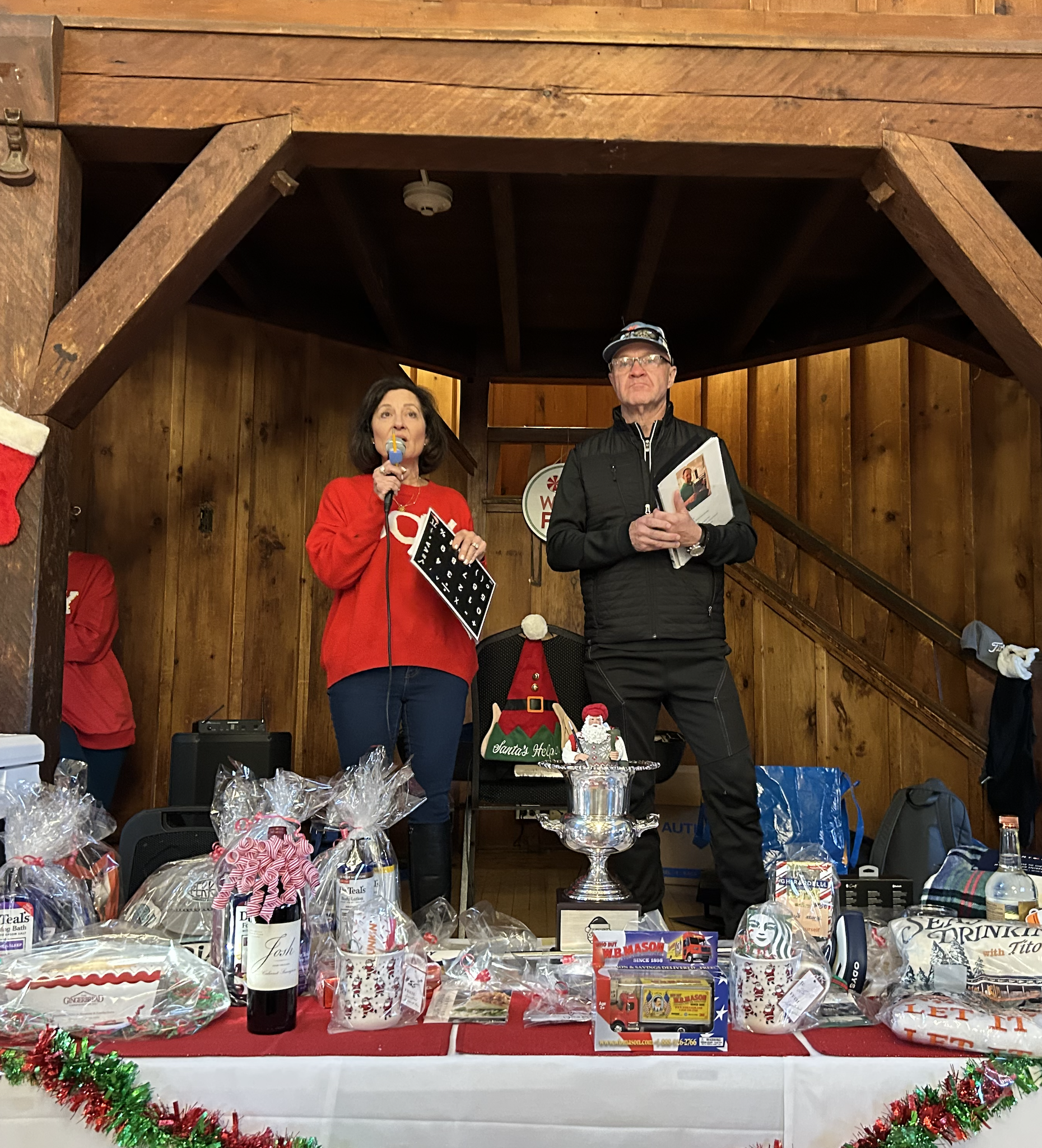 Two people speak at a Christmas charity event beside a table of gift baskets, prizes, and a silver trophy in a rustic wooden room.