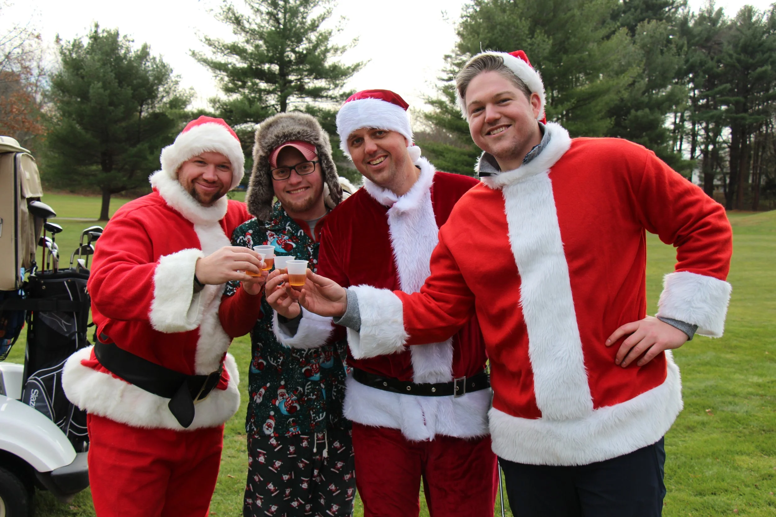 Four men in Santa costumes pose with drinks beside a golf cart on a lawn.