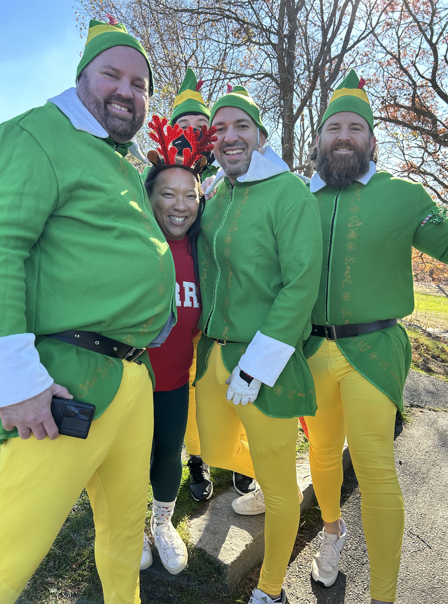 Group of people dressed as Christmas elves, with one person wearing reindeer antlers, posing outdoors.
