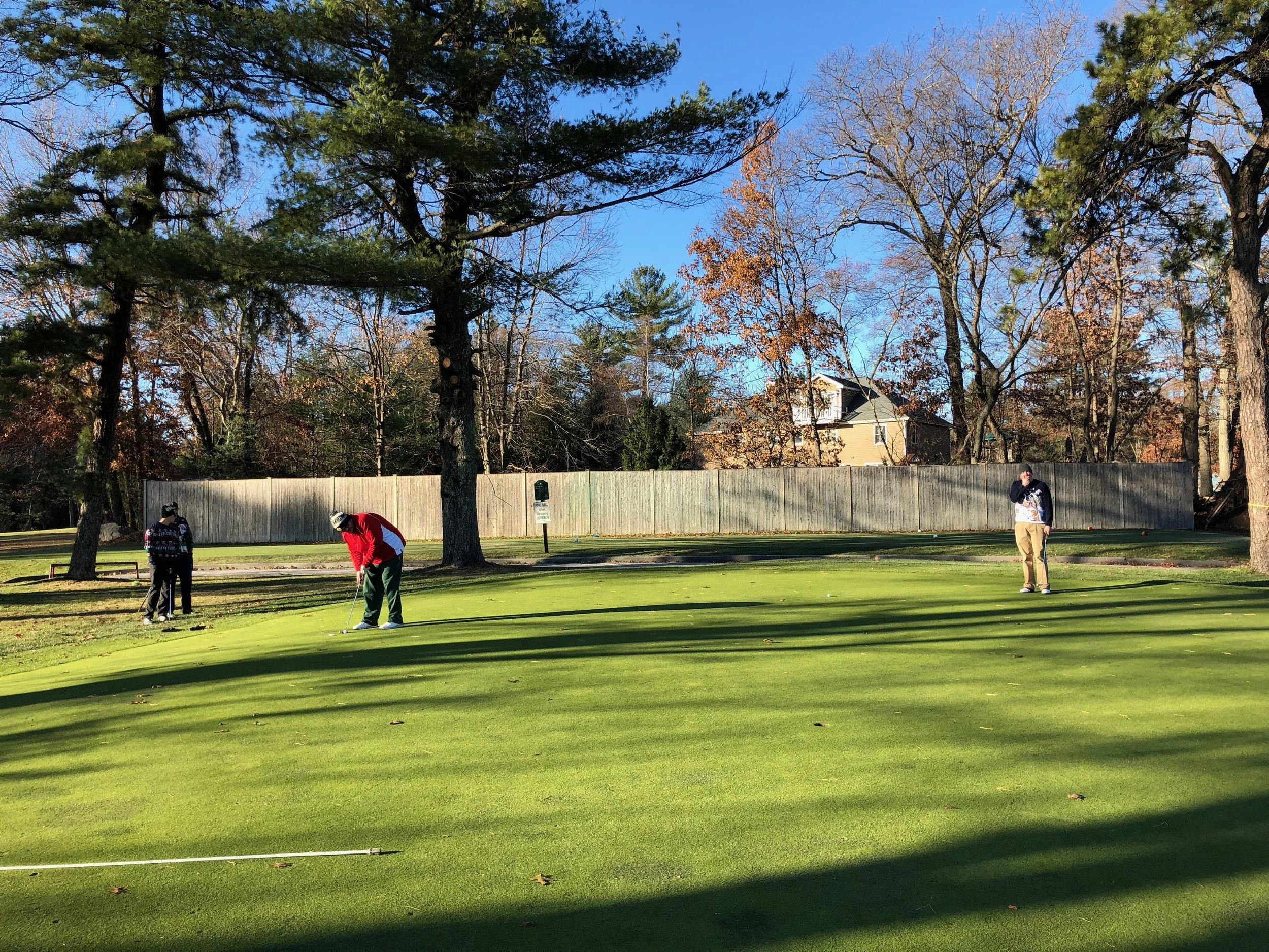 Three golfers playing on a sunny putting green surrounded by trees and a wooden fence.