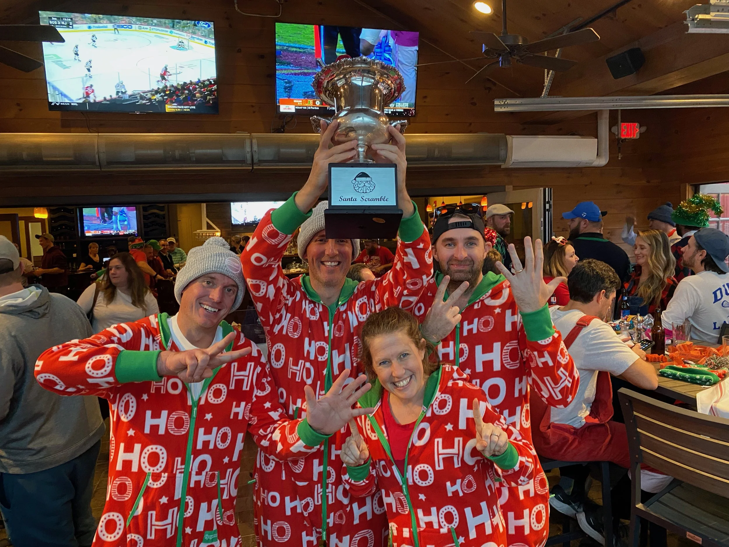 Friends in matching red Christmas pajamas celebrate while holding a trophy at a lively sports bar.