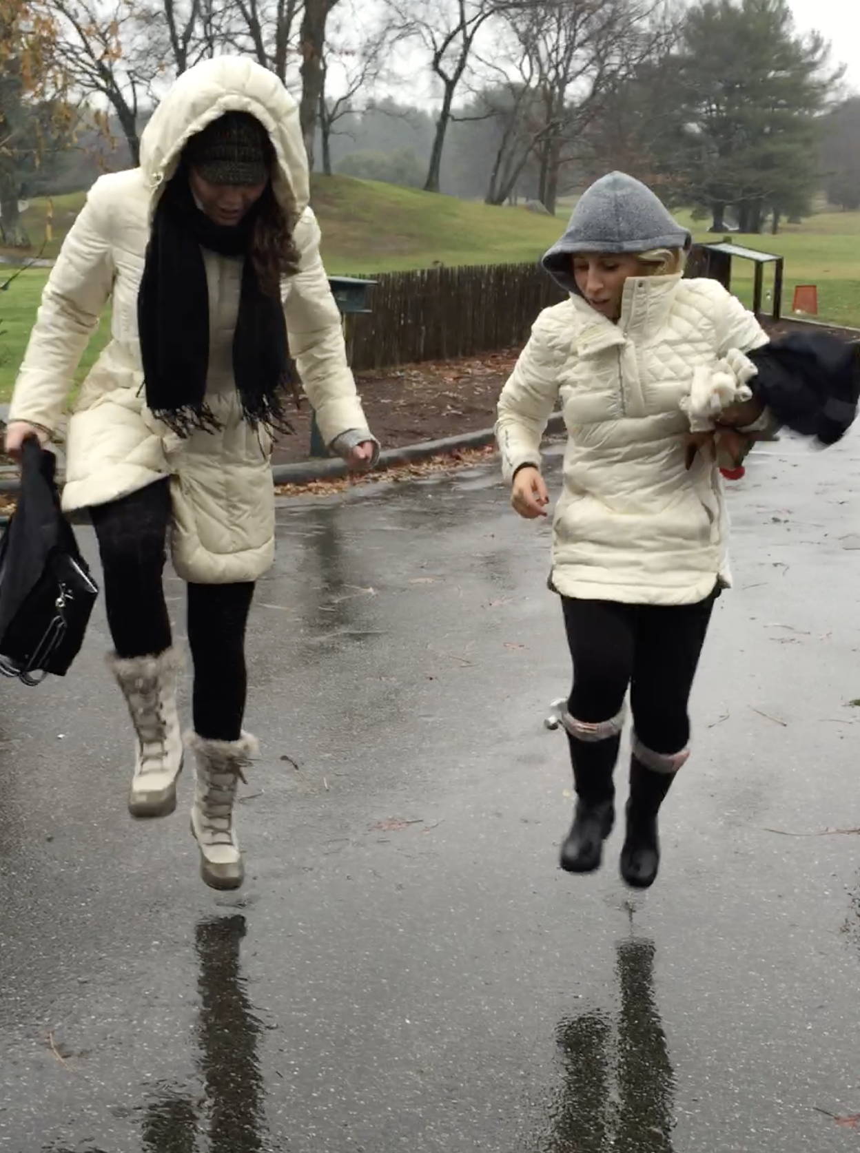 Two women in winter coats jump on a wet park path during a rainy day.
