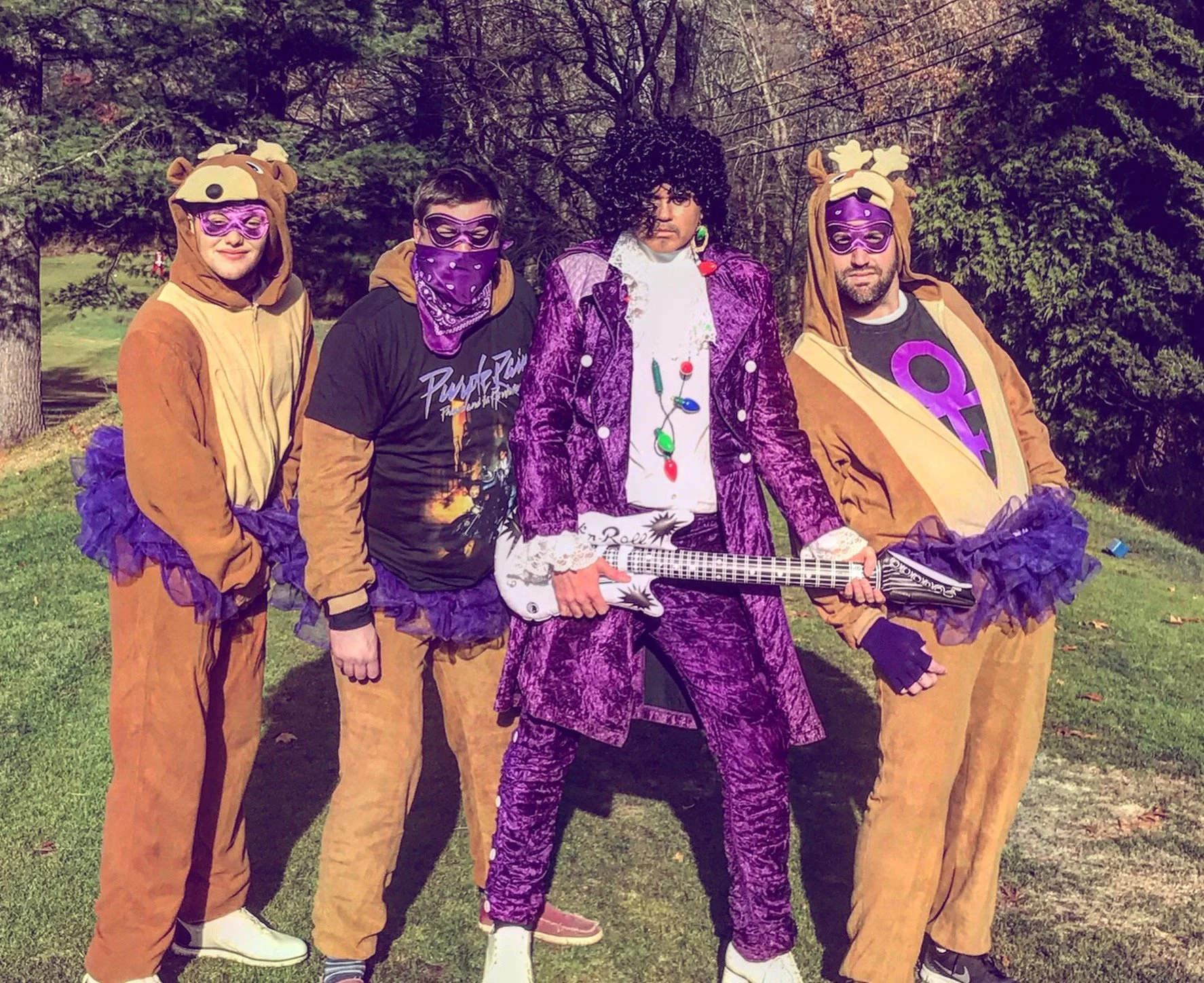 Four people in whimsical purple-and-brown animal costumes pose outdoors, with one holding a white electric guitar.