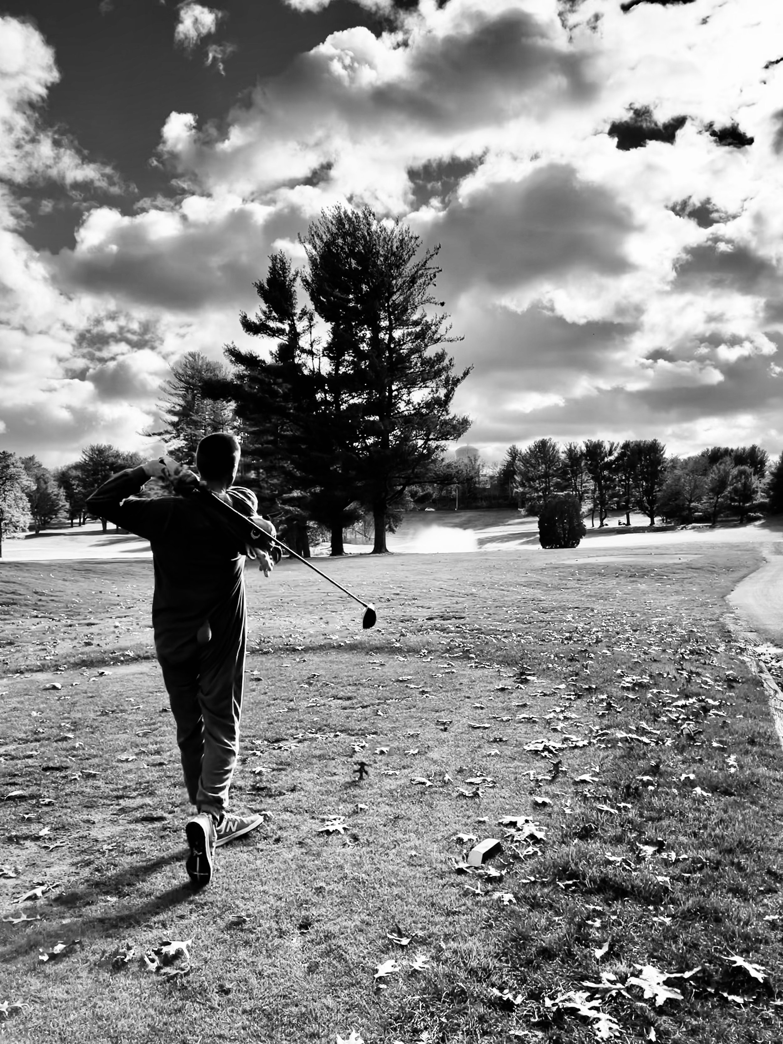 Black-and-white photo of a golfer swinging a club on a tree-lined course beneath dramatic clouds.