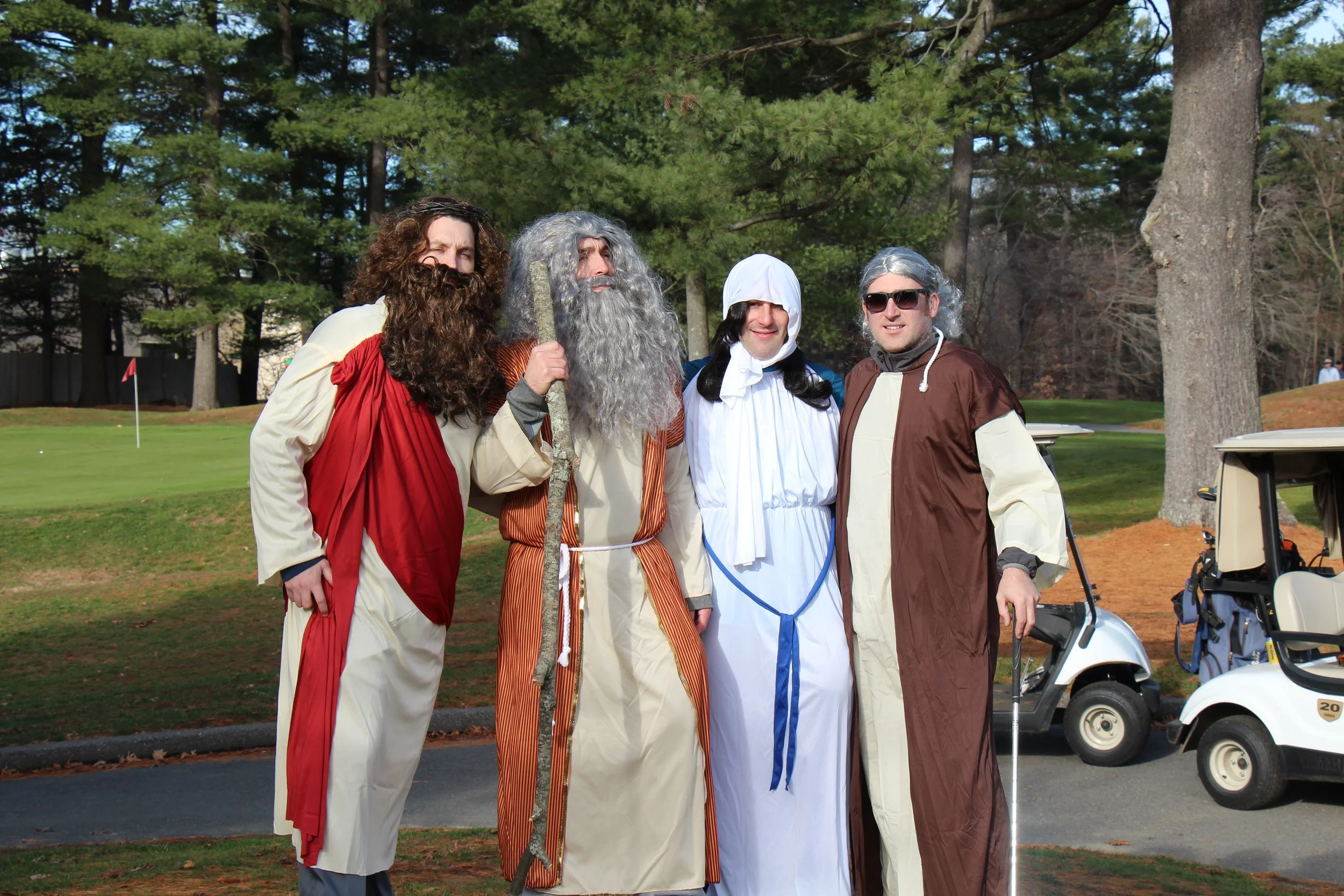 Four people in biblical-era costumes posing outdoors on a golf course, including a bearded man holding a staff and a person wearing sunglasses.