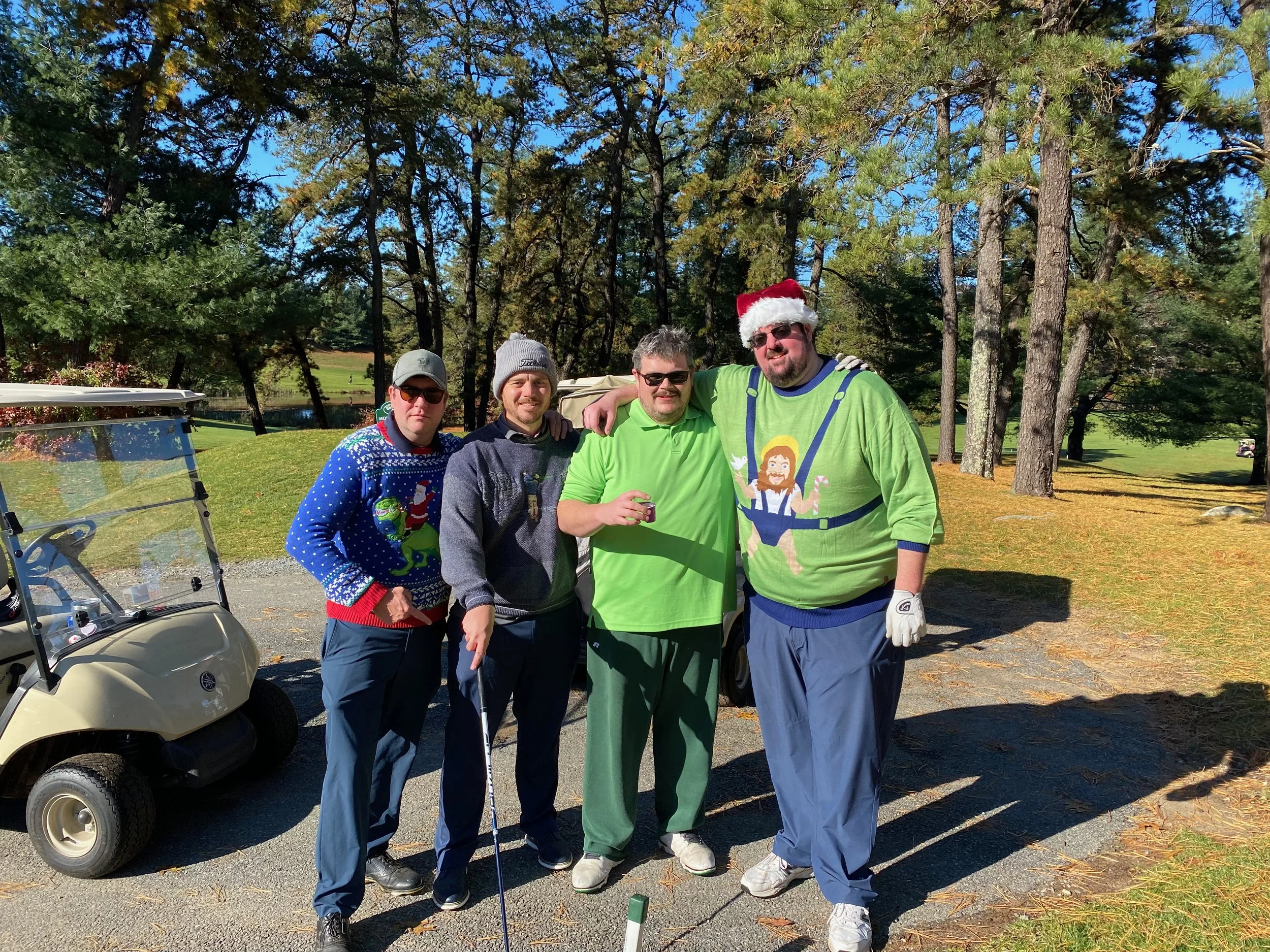 Four men in festive Christmas sweaters pose beside a golf cart on a sunny golf course.