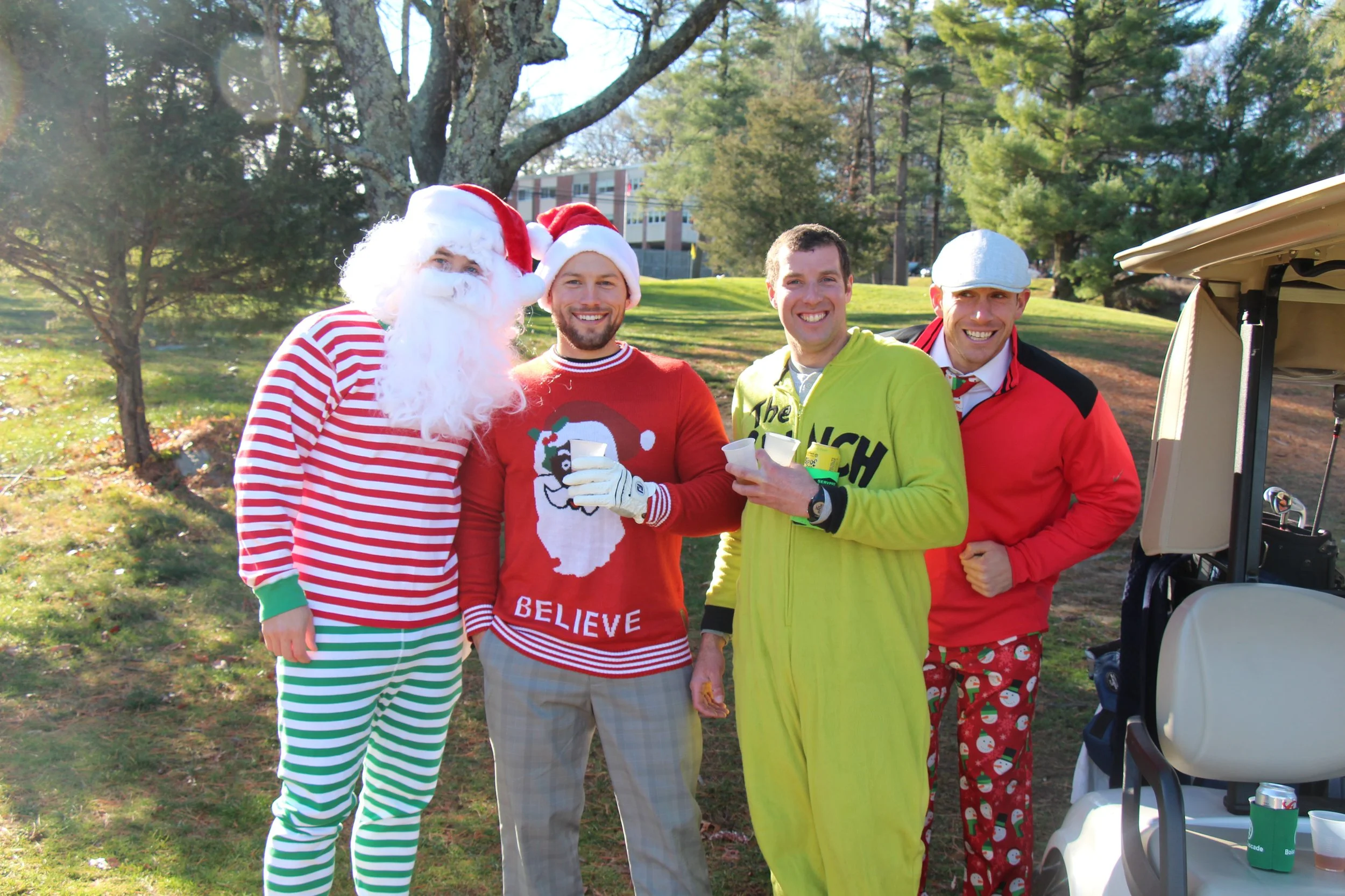 Four men in festive Christmas costumes pose outdoors beside a golf cart, including Santa and an elf.