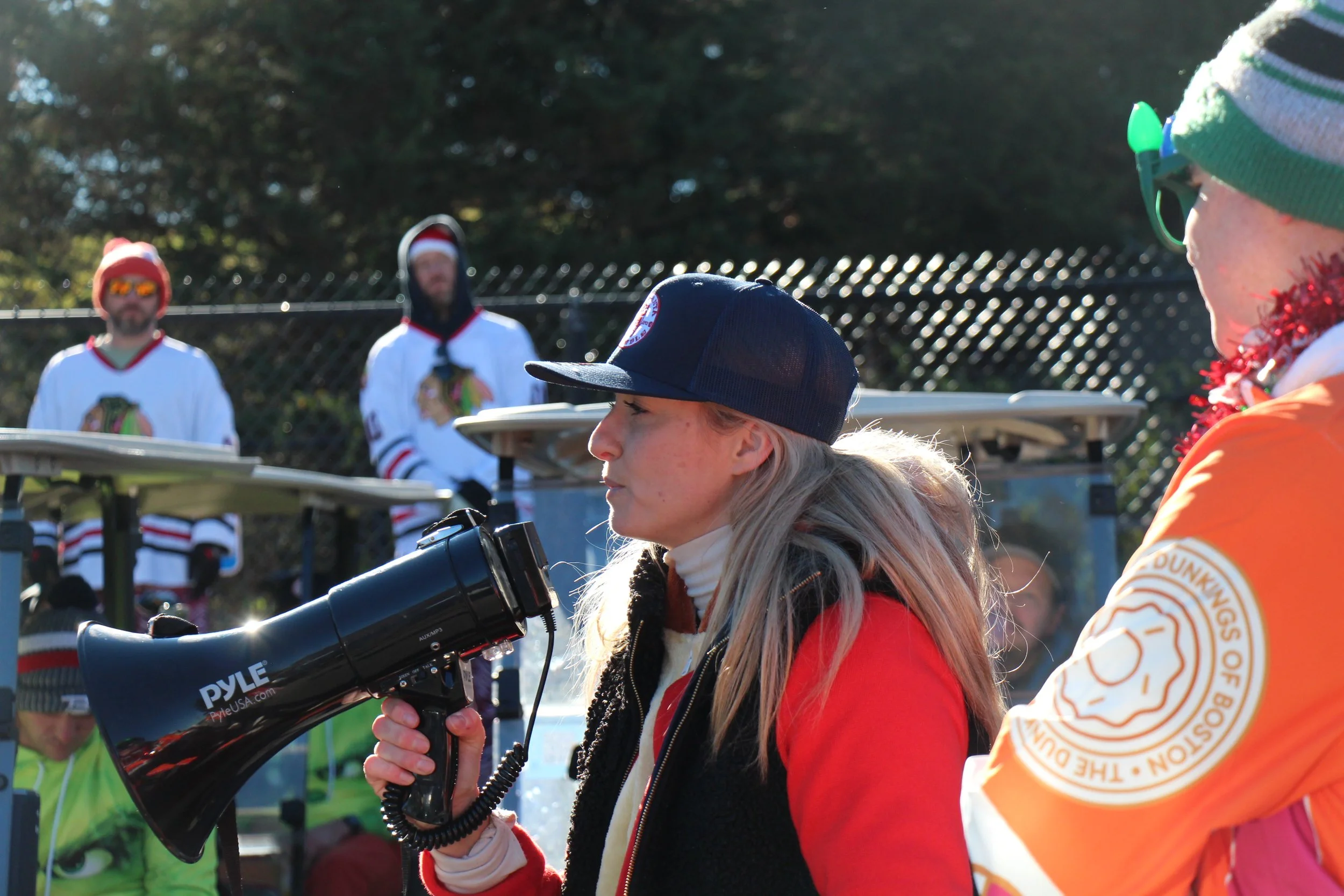 Woman in a black cap holding a megaphone at an outdoor event, with costumed people and a person in an orange shirt in the background.