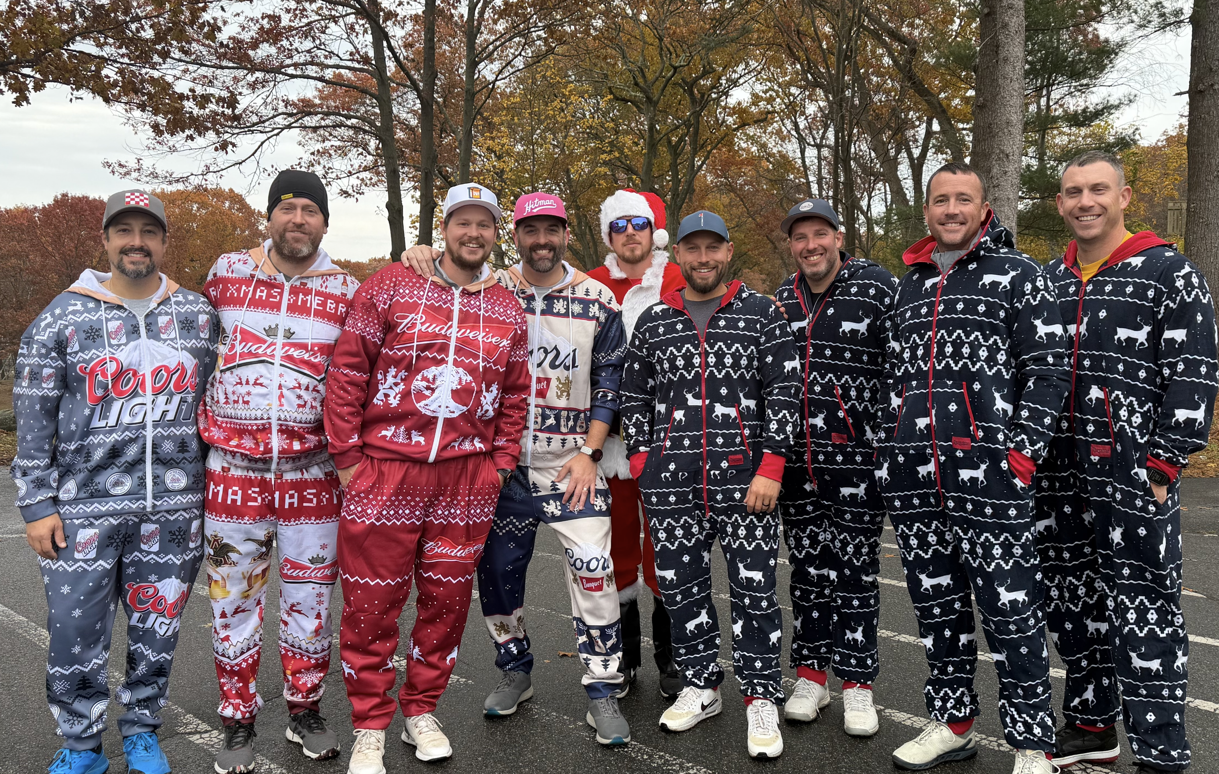 Group of men wearing festive beer-themed Christmas pajamas and one Santa suit posing outdoors among autumn trees.