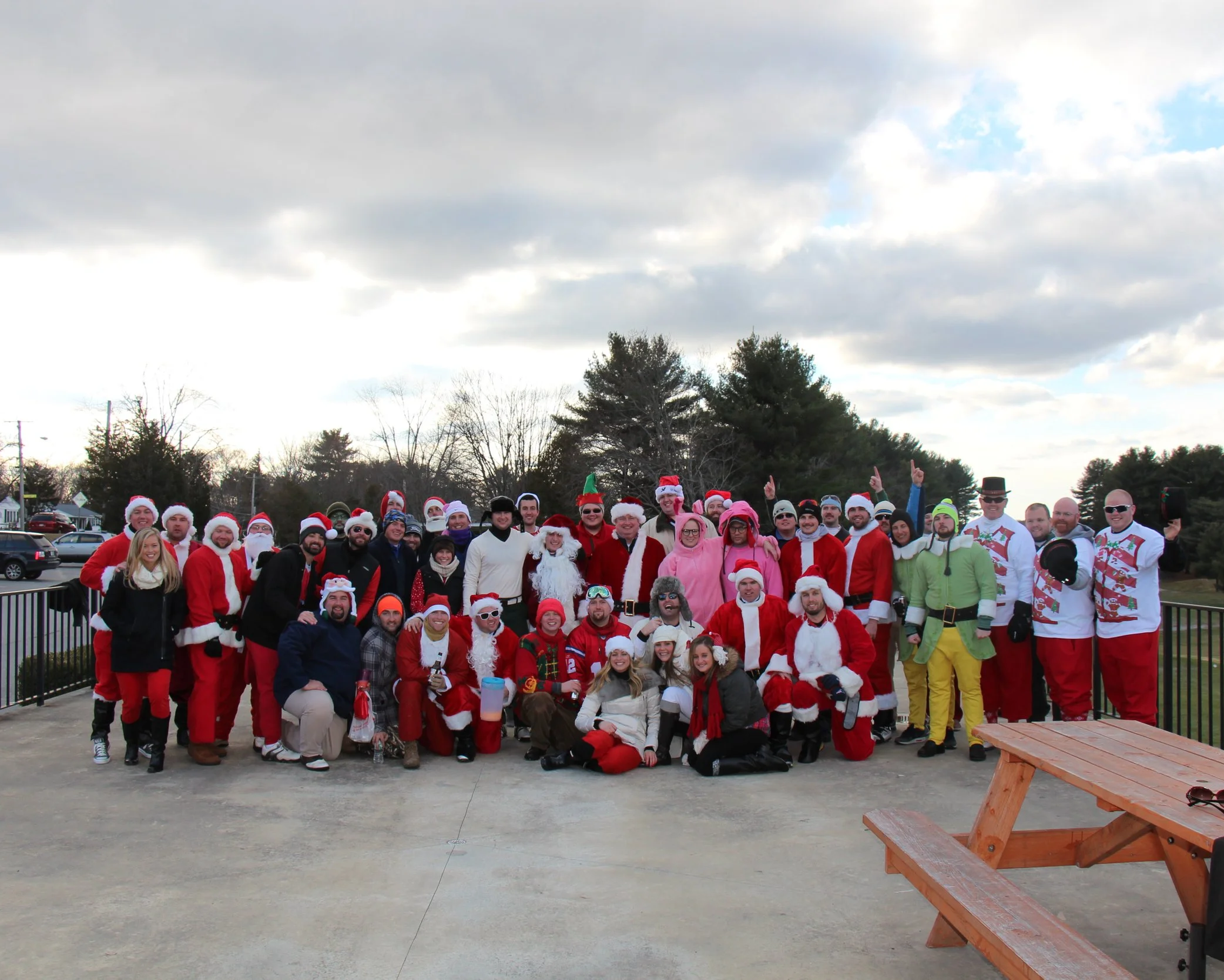 Large group dressed in Santa Claus and other Christmas costumes posing outdoors beside a picnic table