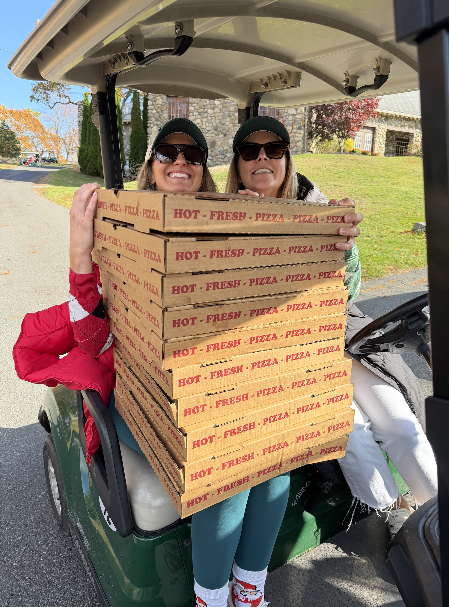 Two women in sunglasses holding a tall stack of hot fresh pizza boxes in a golf cart.