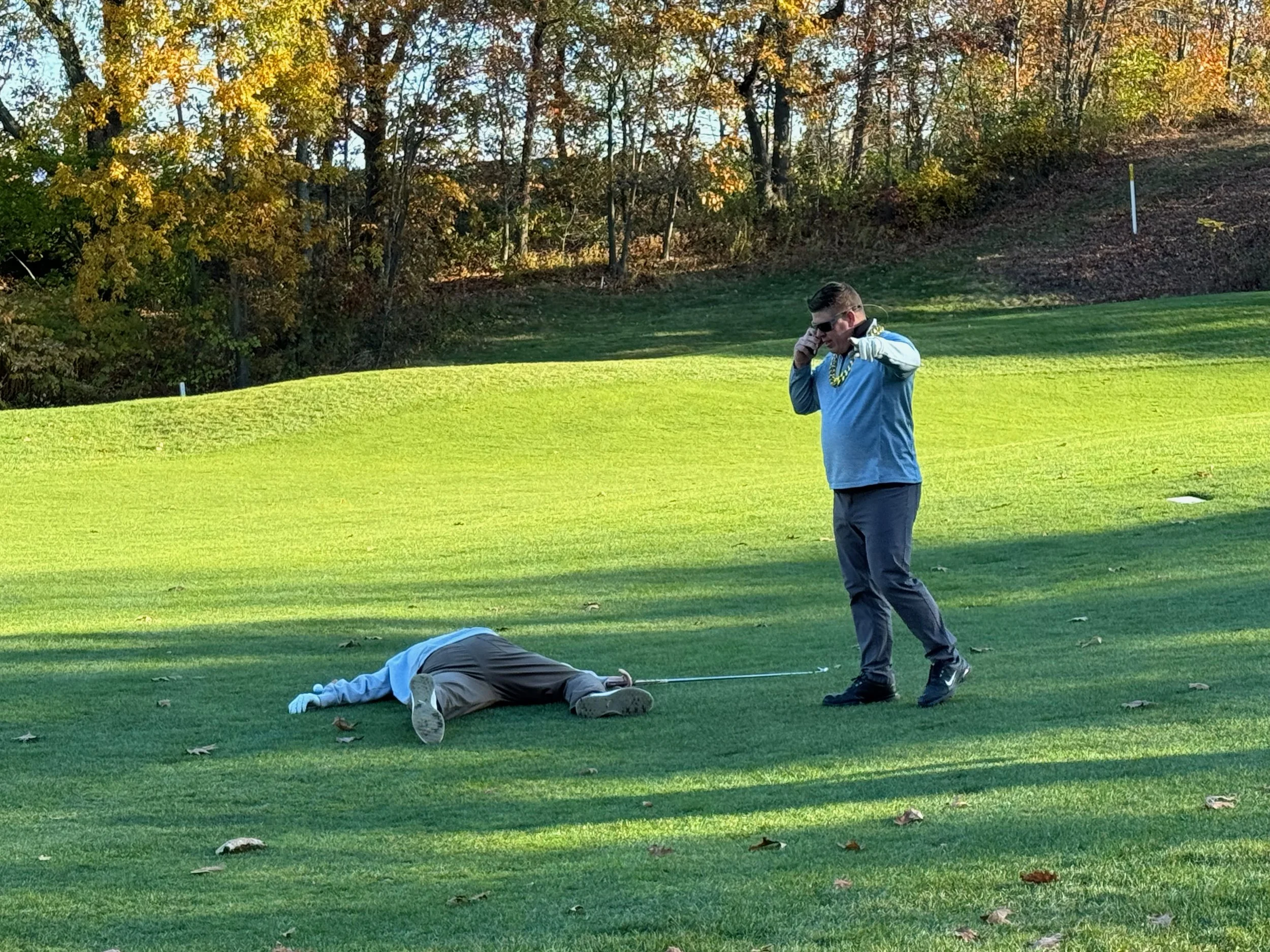 Two golfers on a grassy course, one lying on the ground while another stands beside him holding a phone.