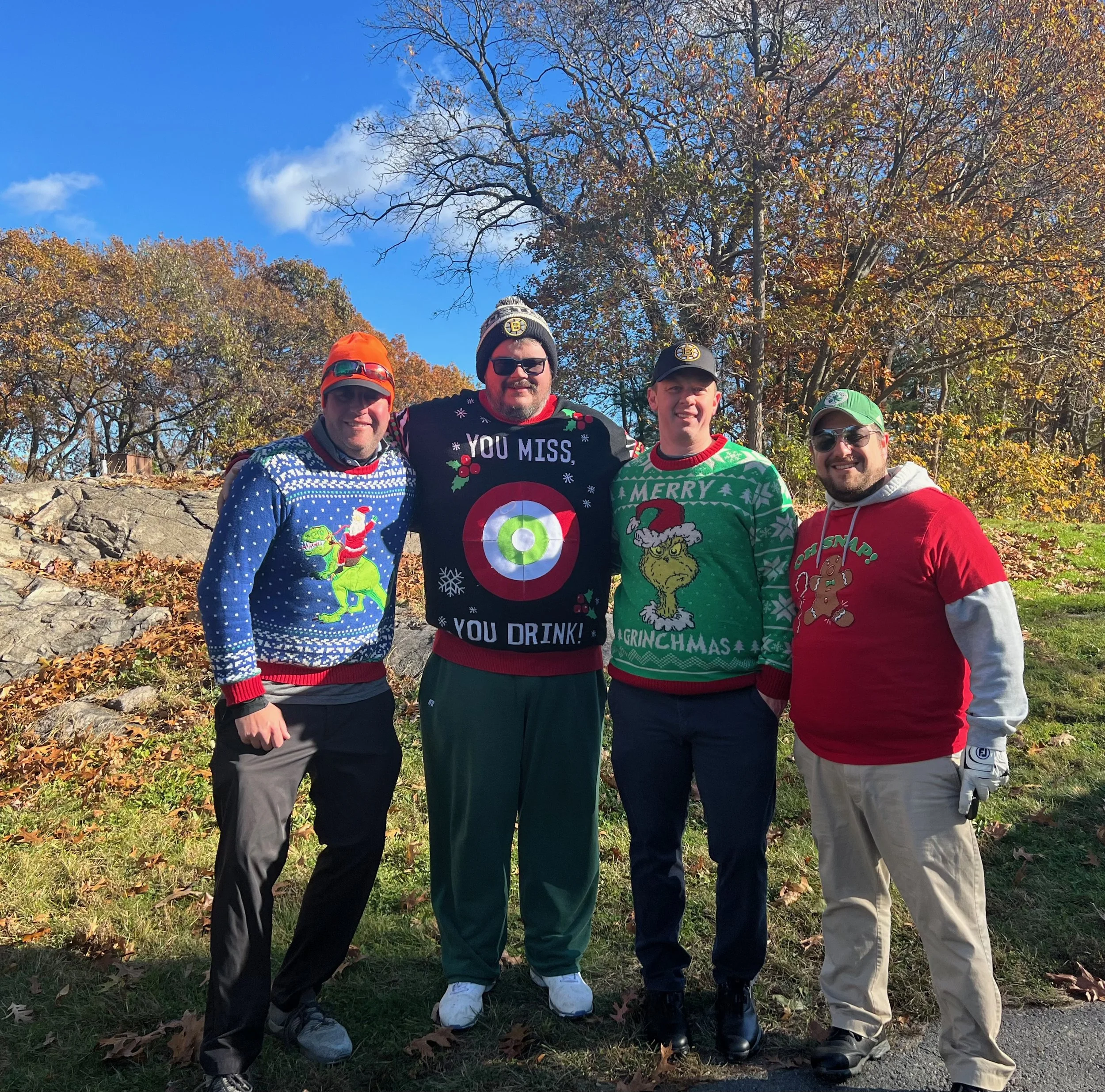 Four men pose outdoors in autumn wearing festive Christmas sweaters and Boston Bruins hats.