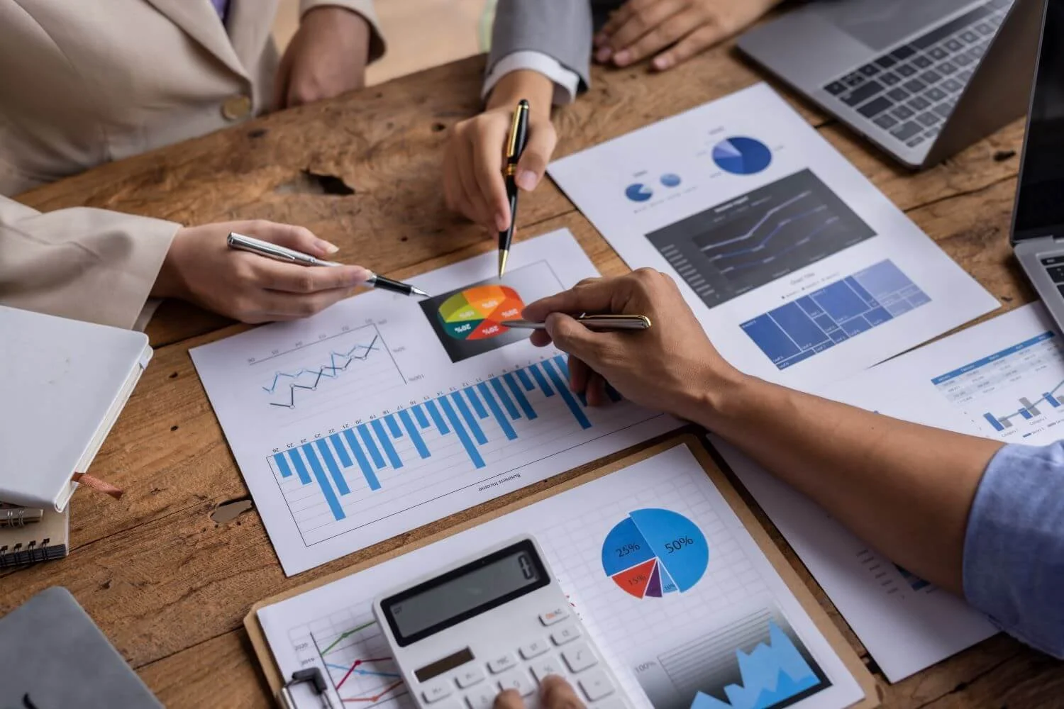 Individuals analyzing and discussing financial charts and graphs on papers, including bar graphs, pie charts, and line graphs, on a wooden table with laptops and a calculator.