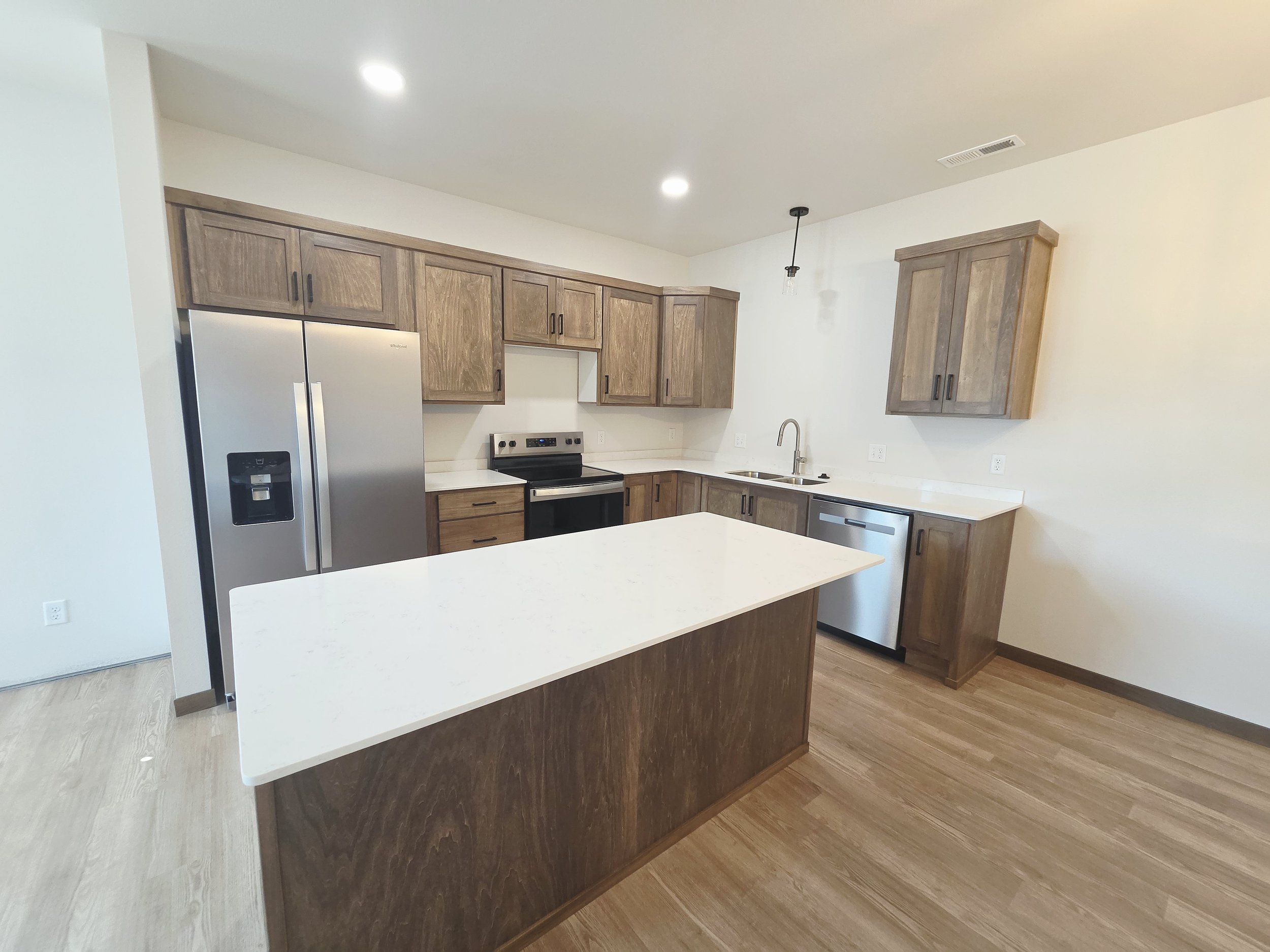 Modern kitchen with wooden cabinets, white countertops, stainless steel appliances including a refrigerator and a dishwasher, and a kitchen island with a white surface.
