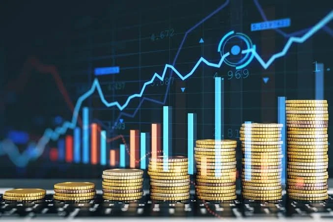Stacks of gold coins increasing in height from left to right, with financial charts and graphs in the background showing upward trends.