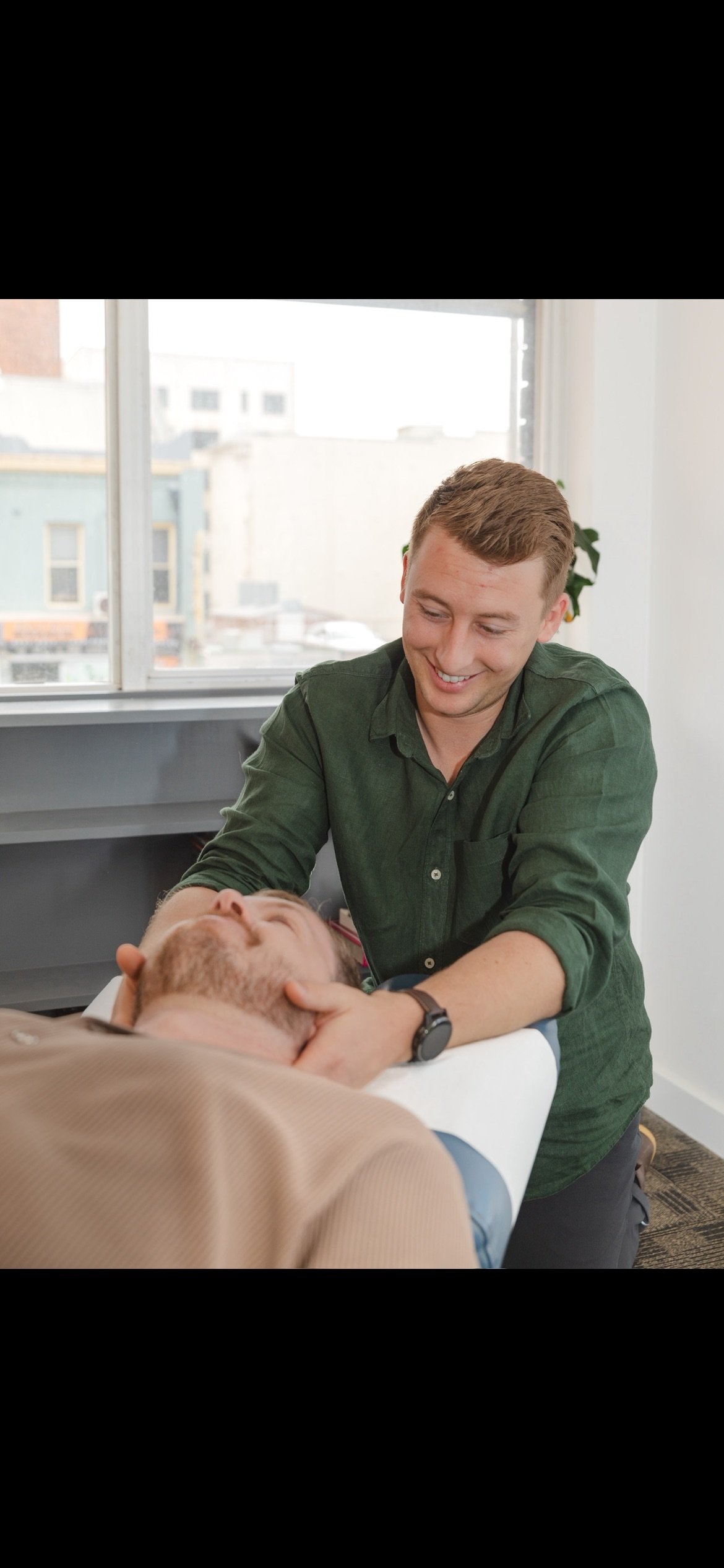 A man with short light brown hair and a dark green shirt is smiling while performing a medical or chiropractic adjustment on another man with a beard, lying on a treatment table. The setting appears to be a medical or chiropractic office with a large window in the background.