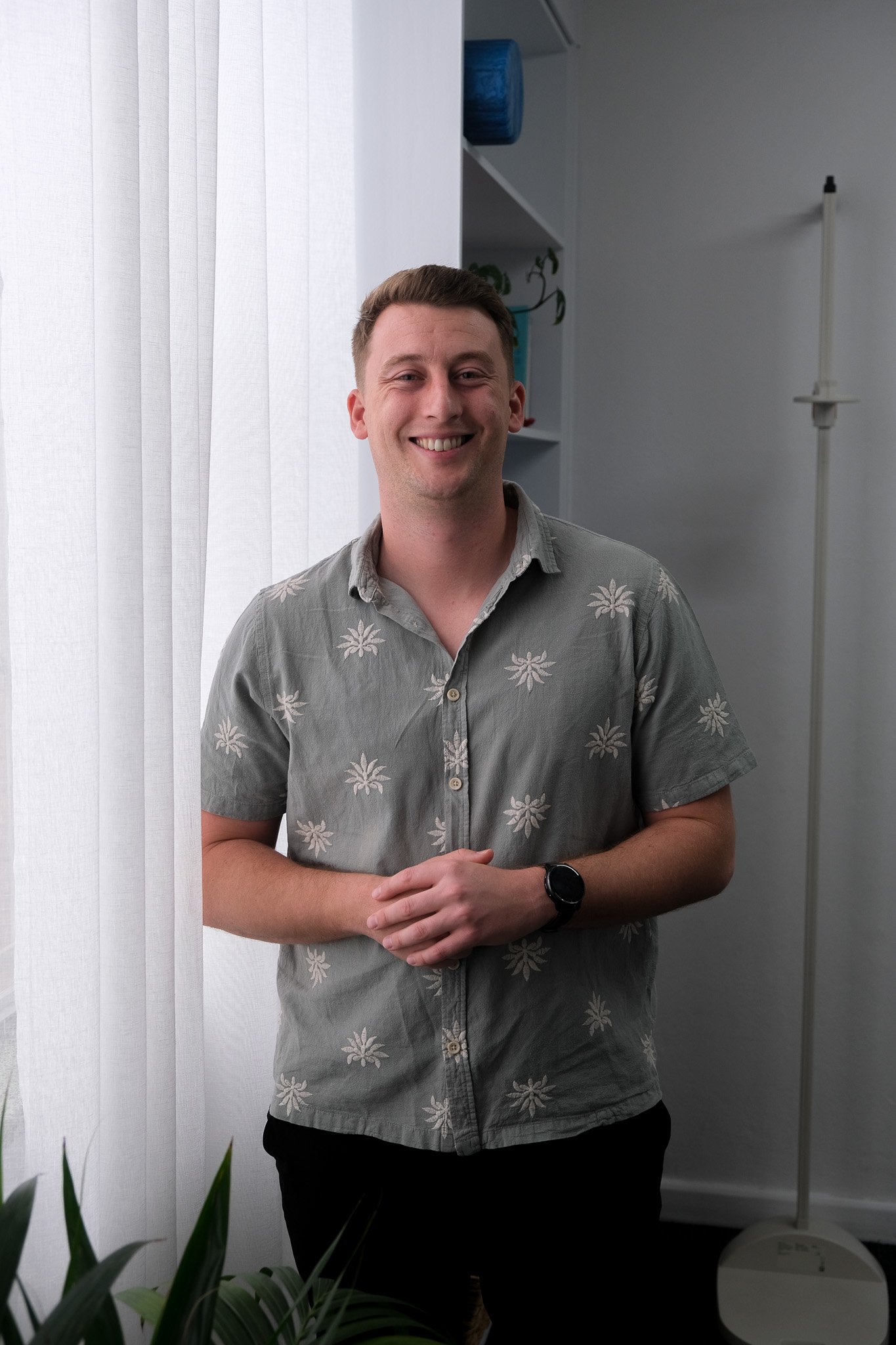 A young man with short brown hair, wearing a gray button-up shirt with white floral patterns, smiling at the camera while standing indoors near a white curtain and a white wall with shelves.