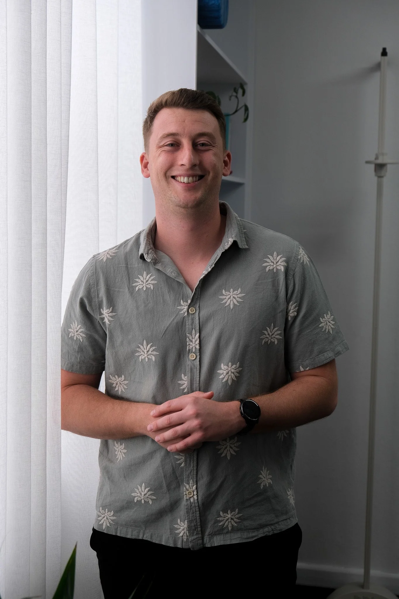 A young man with short brown hair smiling and standing indoors in front of a white curtain and a gray wall, wearing a gray short-sleeved shirt with a white floral pattern, a black watch on his left wrist, and black pants.