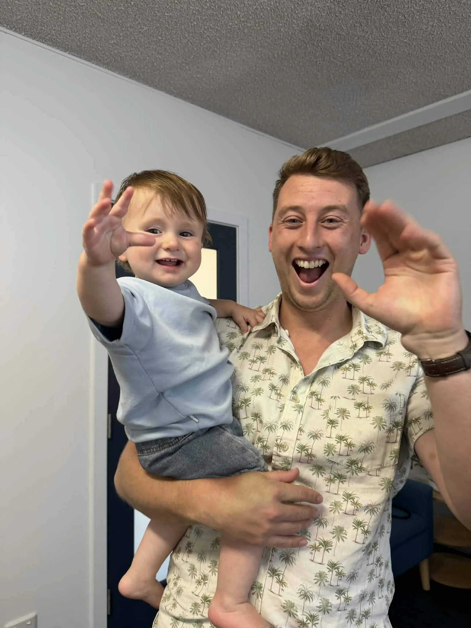 A man smiling and waving while holding a young boy with brown hair and fair skin, who is also smiling and making a peace sign with his hand. The man is wearing a short-sleeved button-up shirt with a palm tree pattern, and the boy is dressed in a light shirt and dark shorts. They are indoors against plain walls.