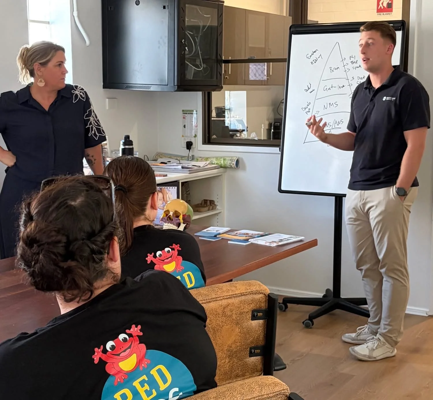 A classroom or meeting room with four people, where two adults are standing at the front and two children are sitting and listening. The woman on the left has blonde hair in a bun, wearing a dark blue dress with floral embroidery on the shoulder, and the man on the right has short brown hair, wearing a black polo shirt and khaki pants, and is writing on a whiteboard. The children are wearing black shirts with a red cartoon character and the word 'RED' on the back. One child has glasses and brown hair tied back. The whiteboard has a diagram with words related to the brain and diet. There is a desk with books and a skull model in front of the children. The room has a window, cabinets, and a fire blanket sign on the wall.