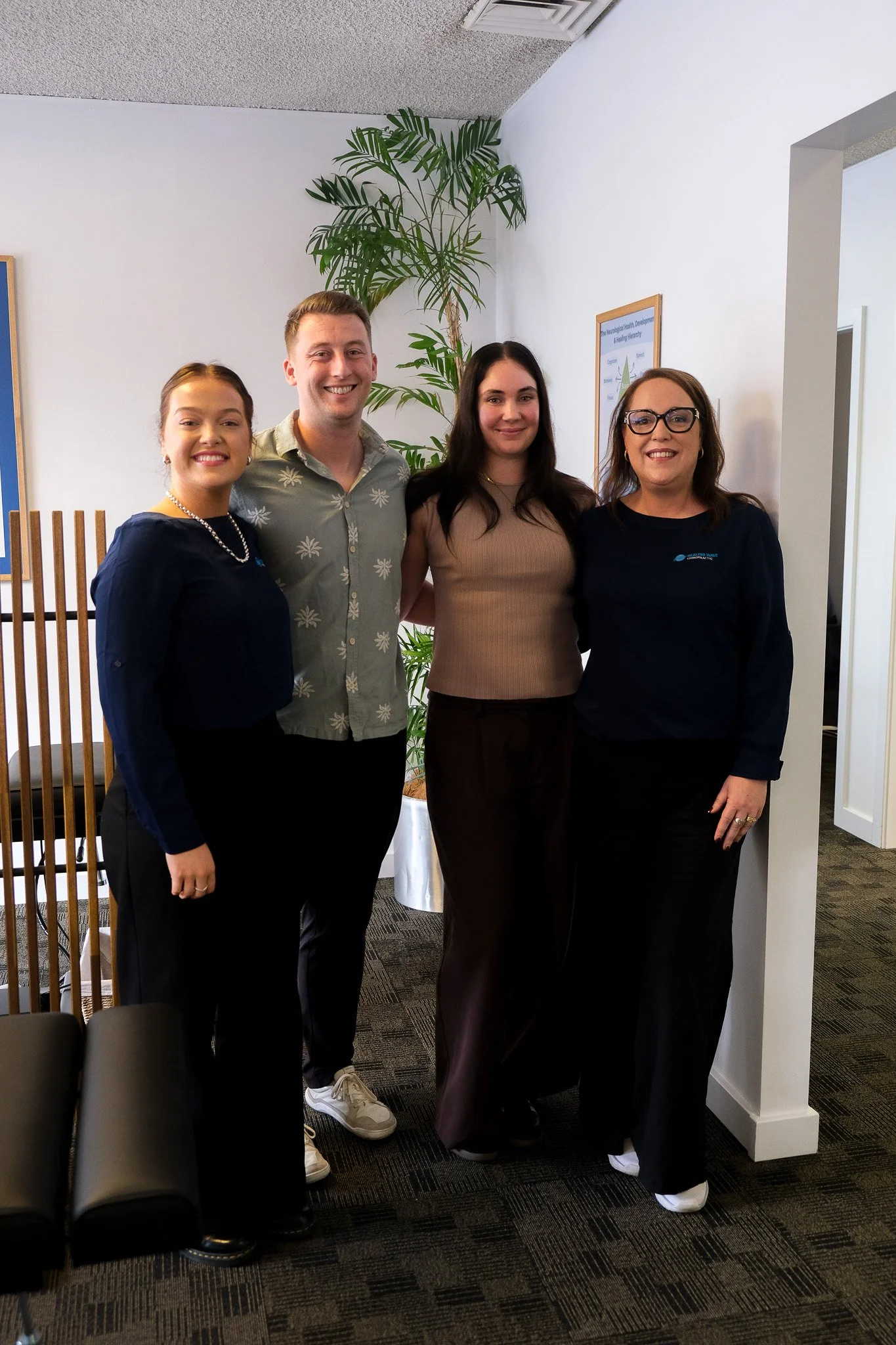 Four smiling people standing closely together indoors, with three women and one man. The woman on the left is wearing a dark blue blouse with a pearl necklace, the man in the middle wears a light green shirt with a floral pattern, the woman next to him is dressed in a brown top, and the woman on the right has on a black top with glasses. A large green plant is visible in the background, along with a framed poster on the wall.