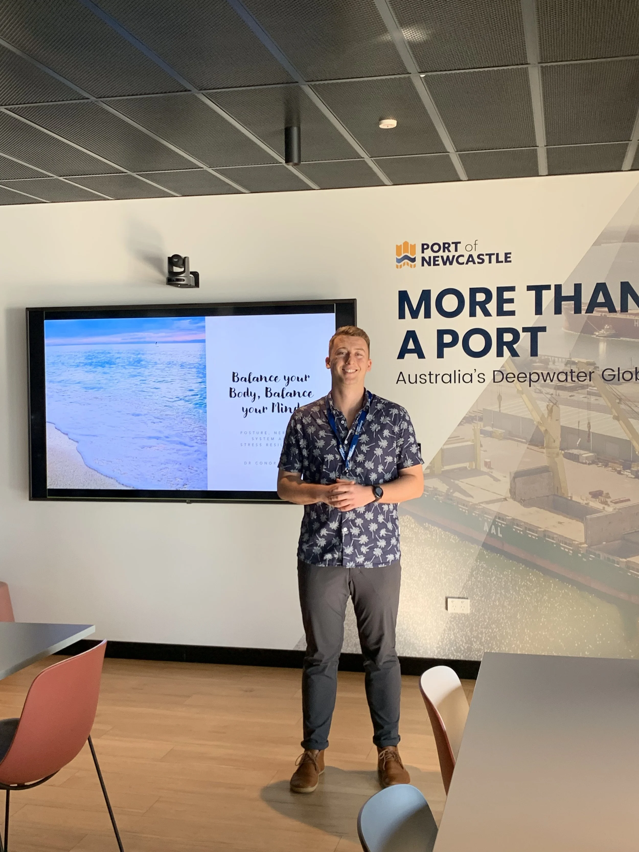 A man is standing in front of a white and blue sign that says "More Than A Port" and "Australia's Deepwater Gateway." There is a large monitor next to him displaying an image of the ocean and text about balancing the body and mind. The man is smiling, wearing a floral shirt, gray pants, and brown shoes, with a watch on his left wrist and a lanyard around his neck.