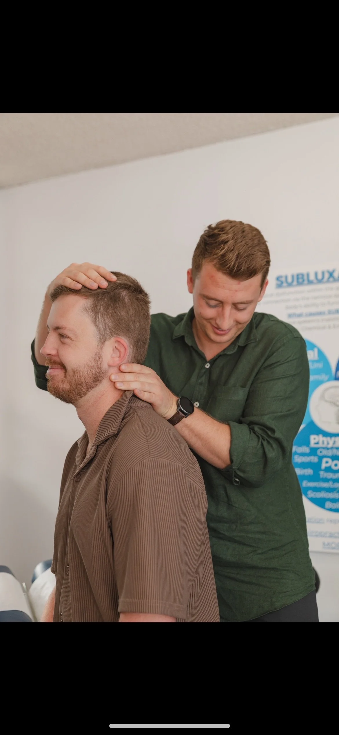 A chiropractor adjusts a man's neck during a session, both smiling and in a clinical setting with a white wall and a poster in the background.