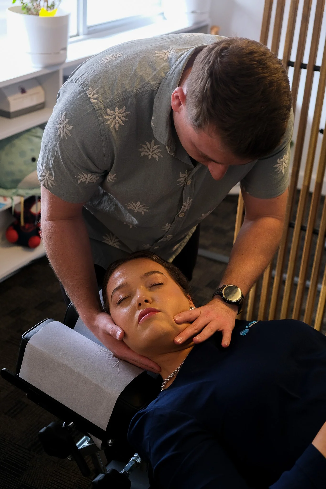 A woman lying on a chiropractic table receiving neck adjustment from a male chiropractor in a treatment room, with a window and potted plant in the background.