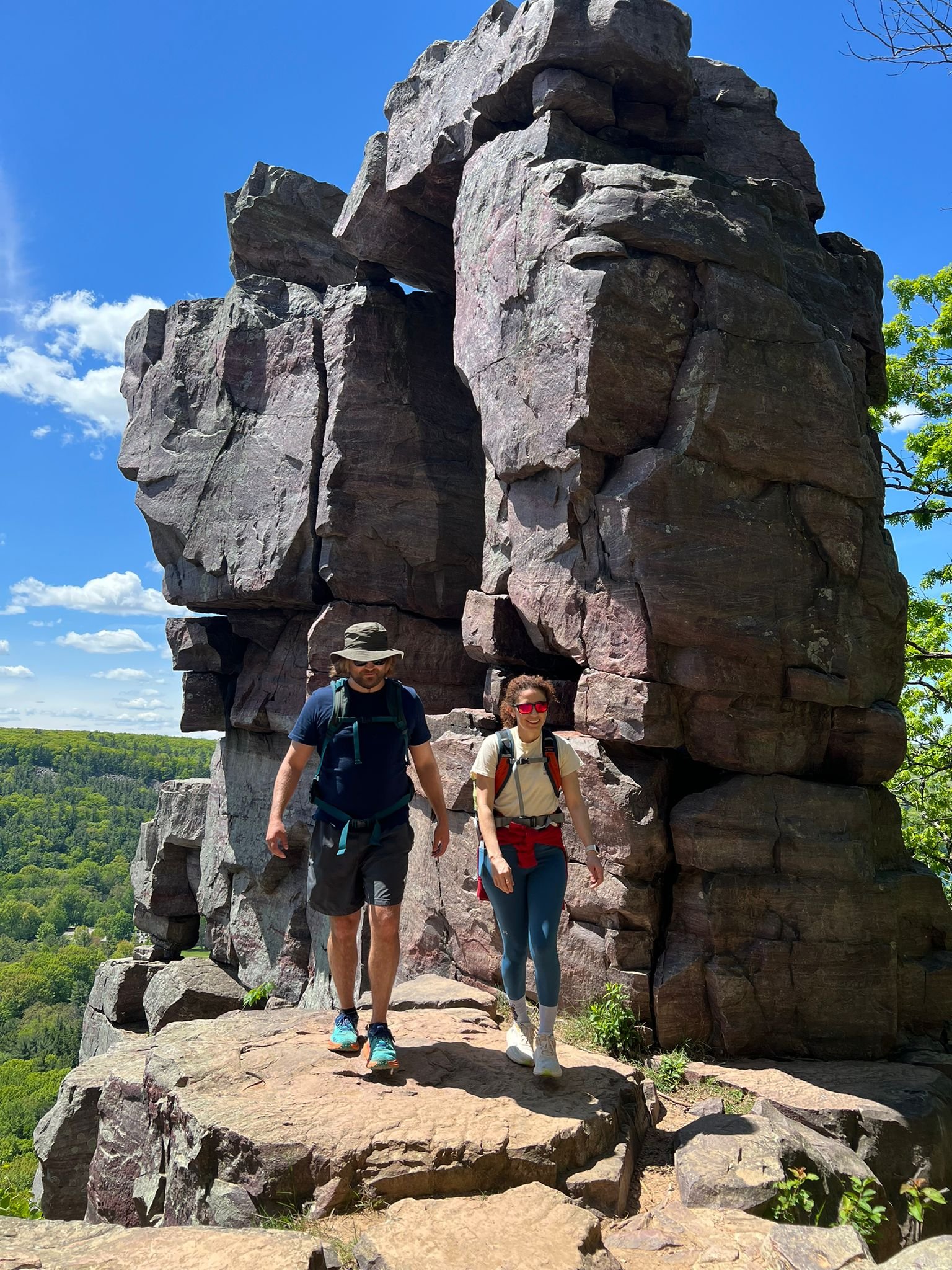 Two hikers walk down a rocky ledge in front of a large, natural rock formation that has a hole or arch in the middle. The sky is clear and blue with a few clouds, and greenery surrounds the area.