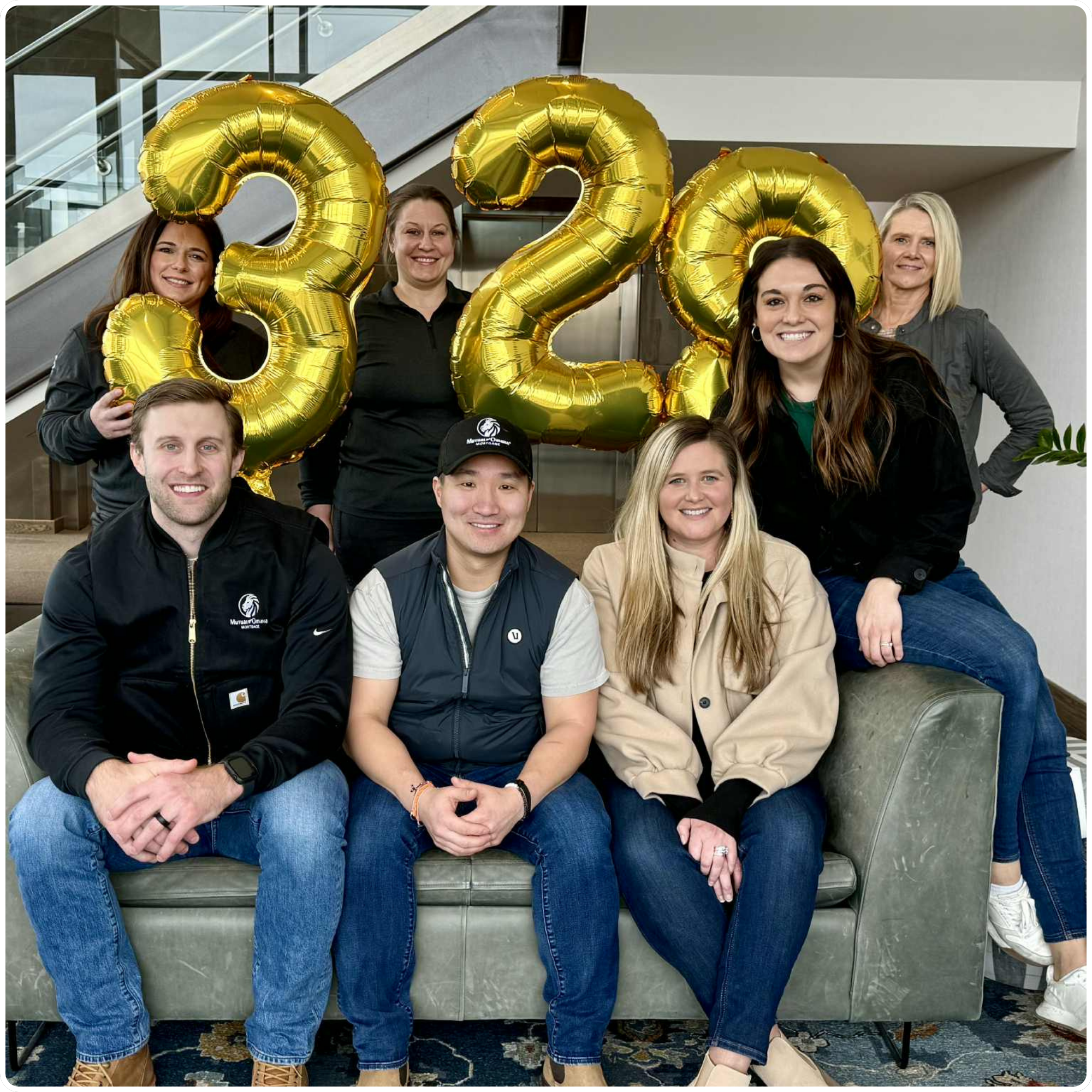 Group of eight coworkers posing with large gold number balloons in a modern office lounge.