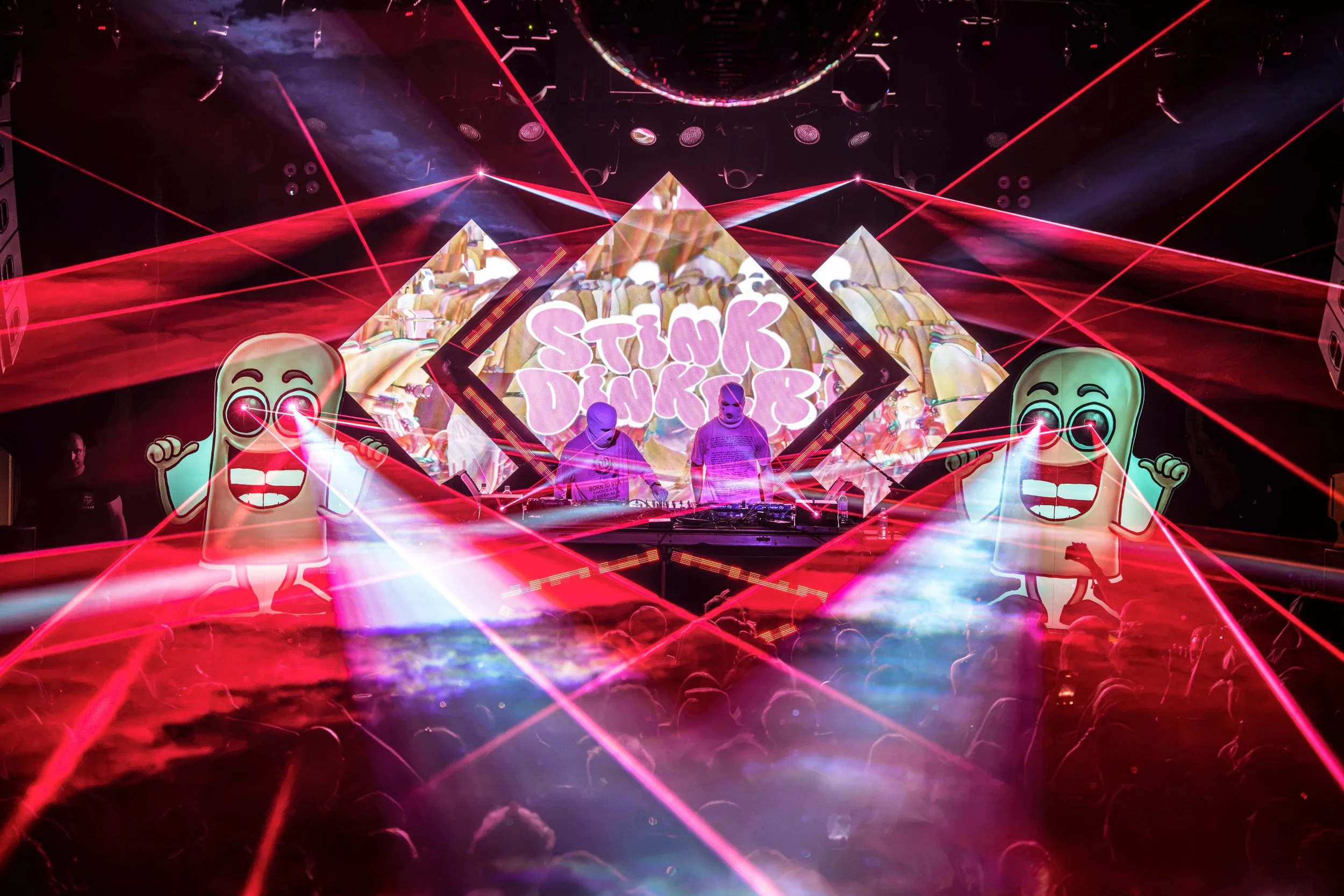 A vibrant scene with laser lights, cartoonish characters, and the words 'STINK DINKER' displayed in bright pink on a large digital screen, creating an energetic nightclub or concert atmosphere.