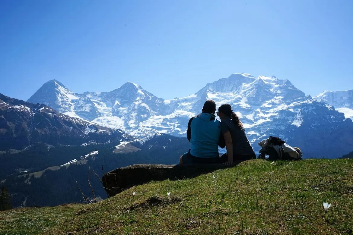 A couple sitting on a grassy hilltop with snow-capped mountains in the background.