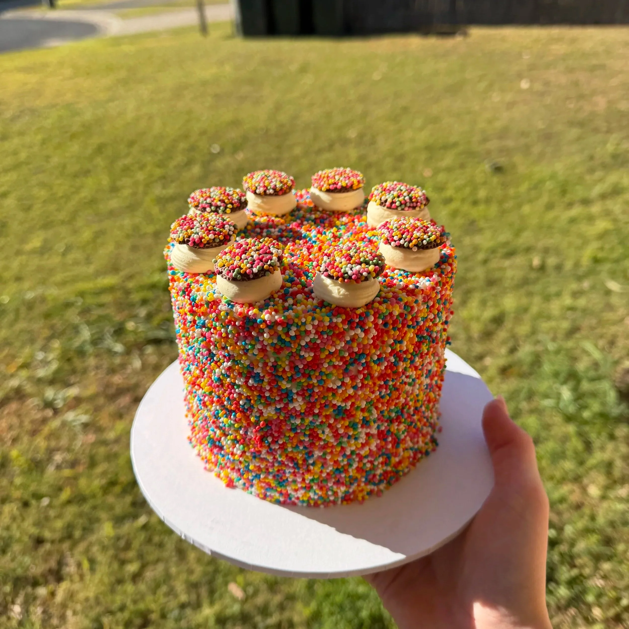 A cake decorated with multicolored sprinkles on top and around the sides, topped with small buttercream drops with rainbow sprinkles.