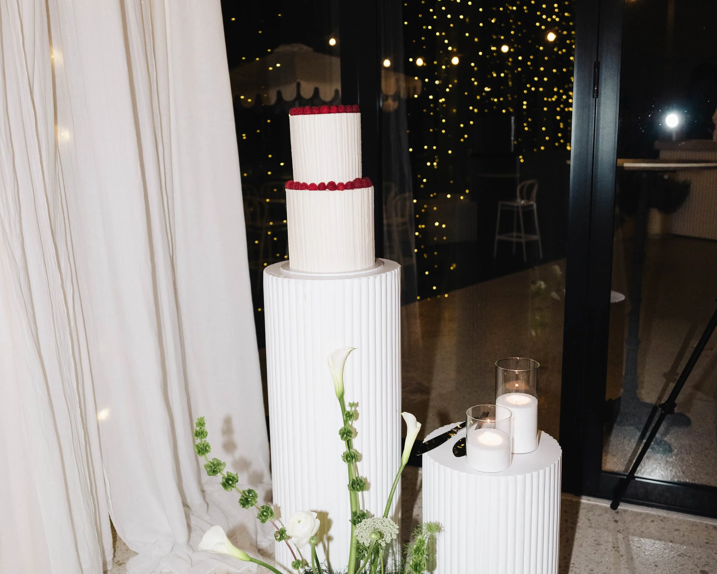 Tall tiered wedding cake decorated with white vertical lines and red berries on top, surrounded by candles and floral arrangements, with a backdrop of string lights outside.