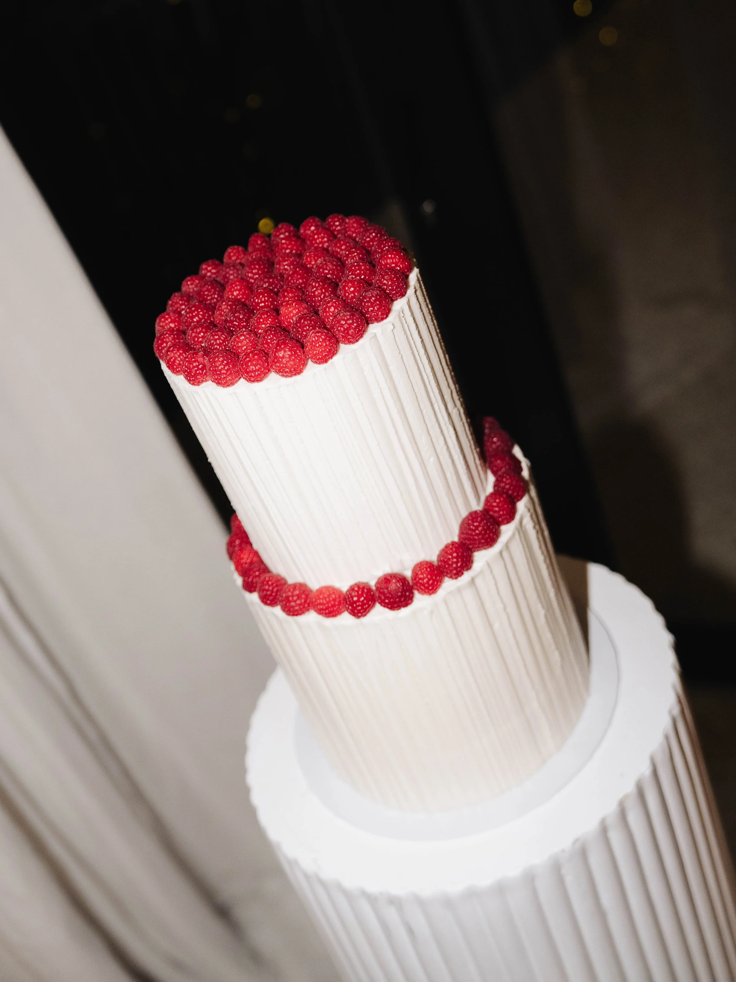 A tall, white, textured cake decorated with a border of red raspberries on top and around the middle, placed on a white cake board.