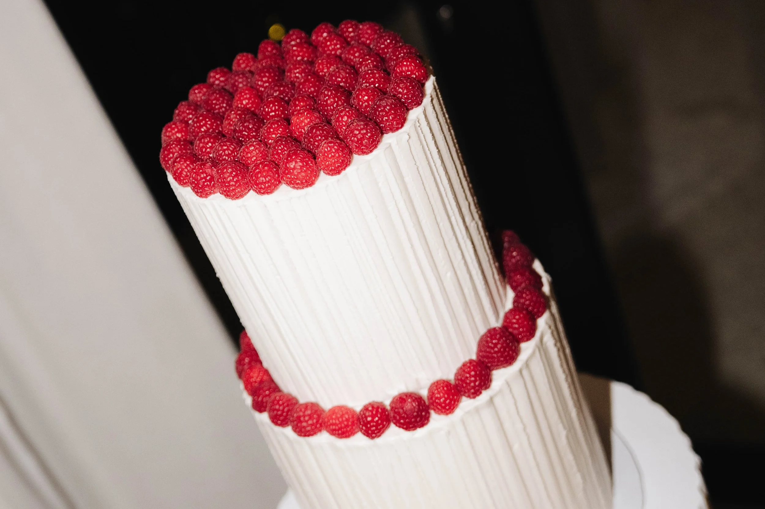 A tall, white, tiered cake decorated with fresh raspberries along the top and around the sides, with a black background.