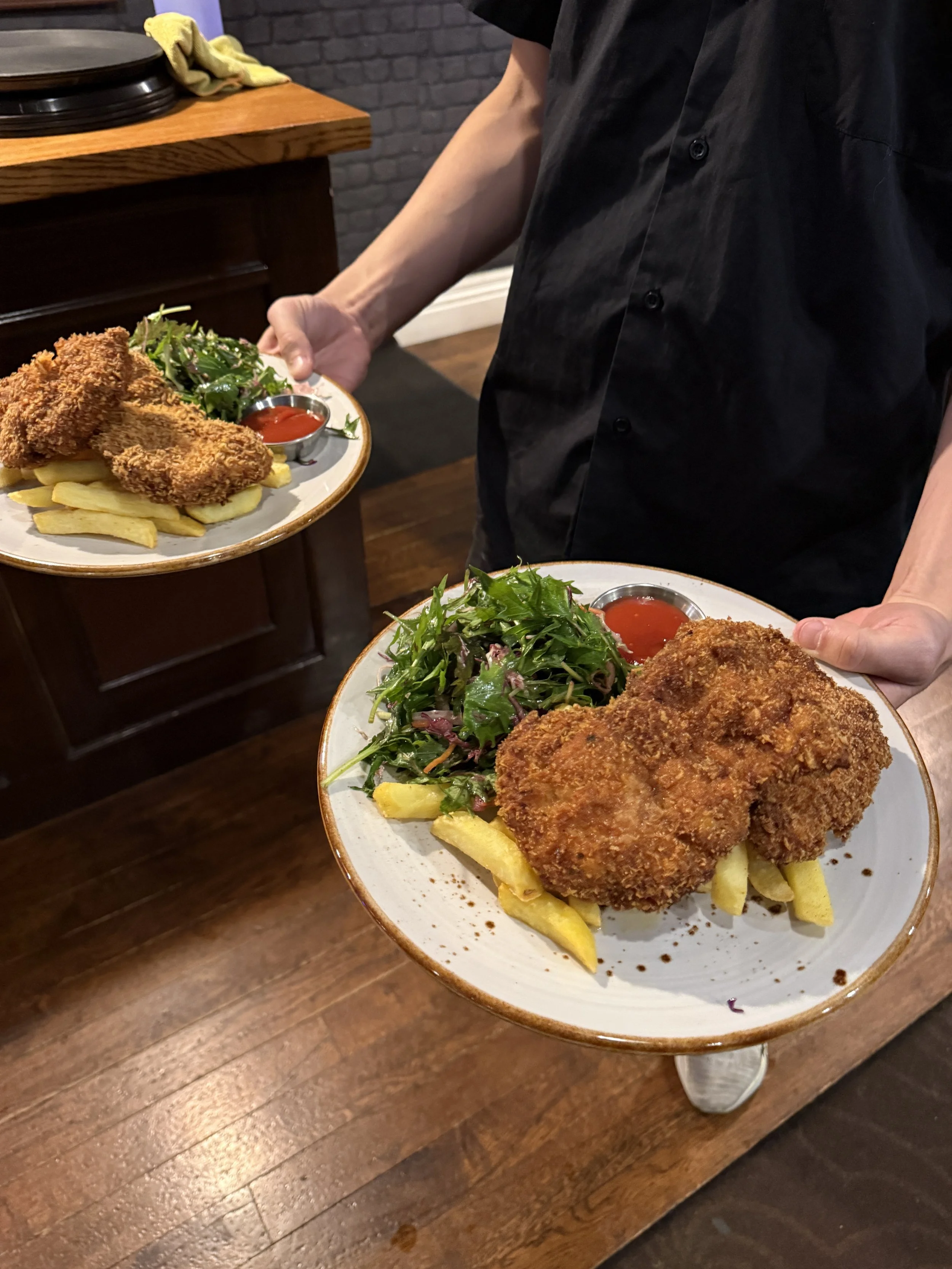 A person holding a plate with fried chicken, French fries, and a side salad with mixed greens, served with a cup of ketchup.