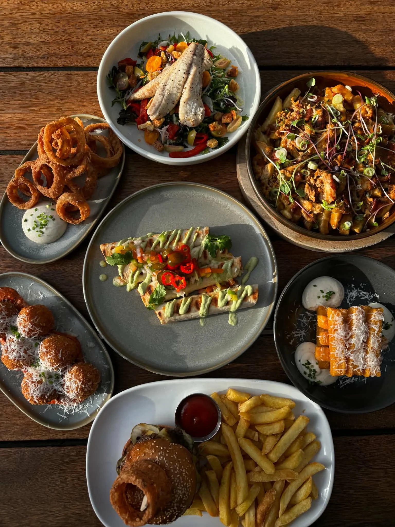 Assorted fast food dishes on a wooden table, including onion rings, chicken salad, pizza, a burger with fries, and a dessert with coconut flakes.