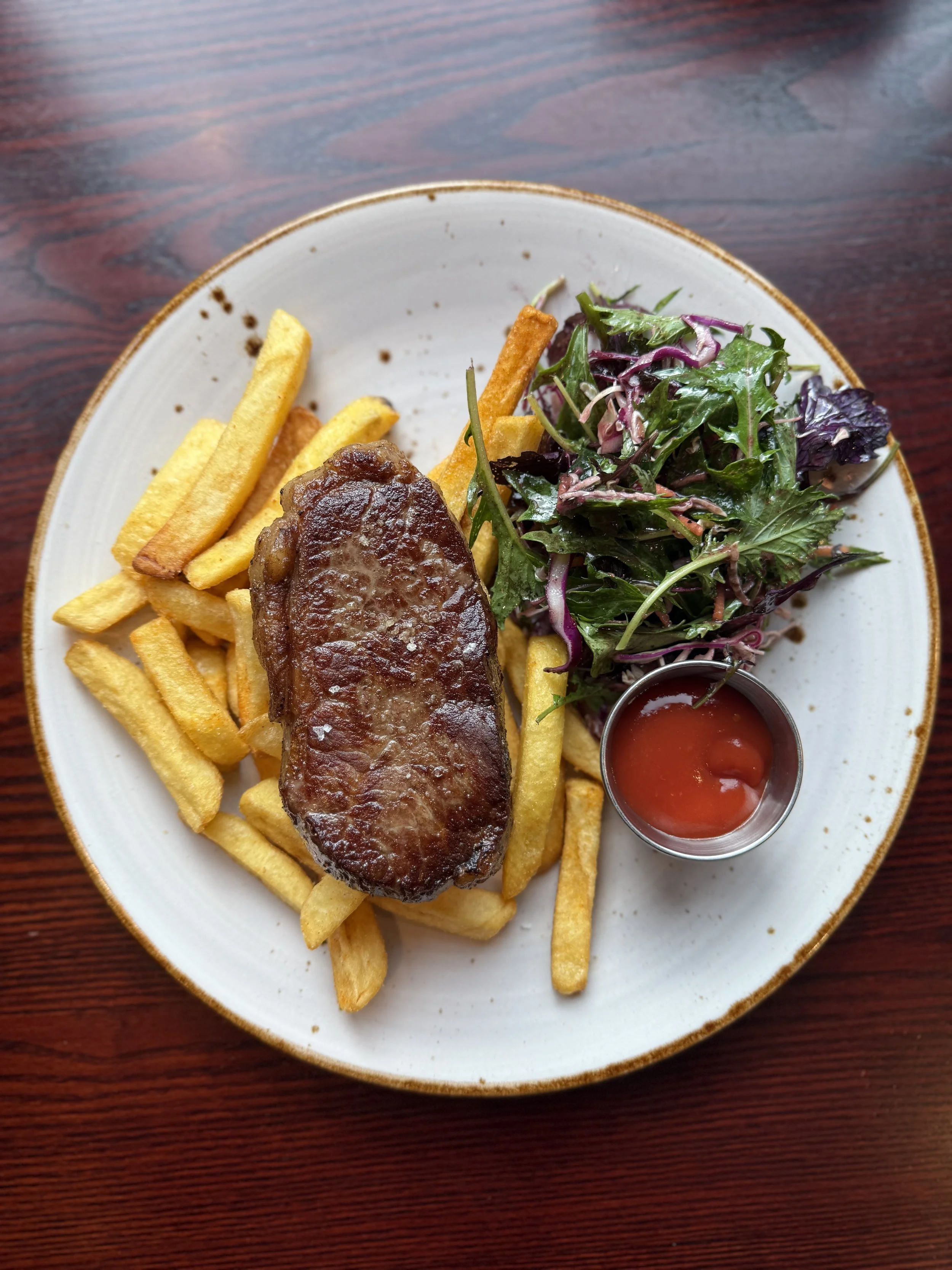 Plate of French fries, a cooked steak, a side salad with mixed greens and purple cabbage, and a small cup of ketchup on a wooden table.