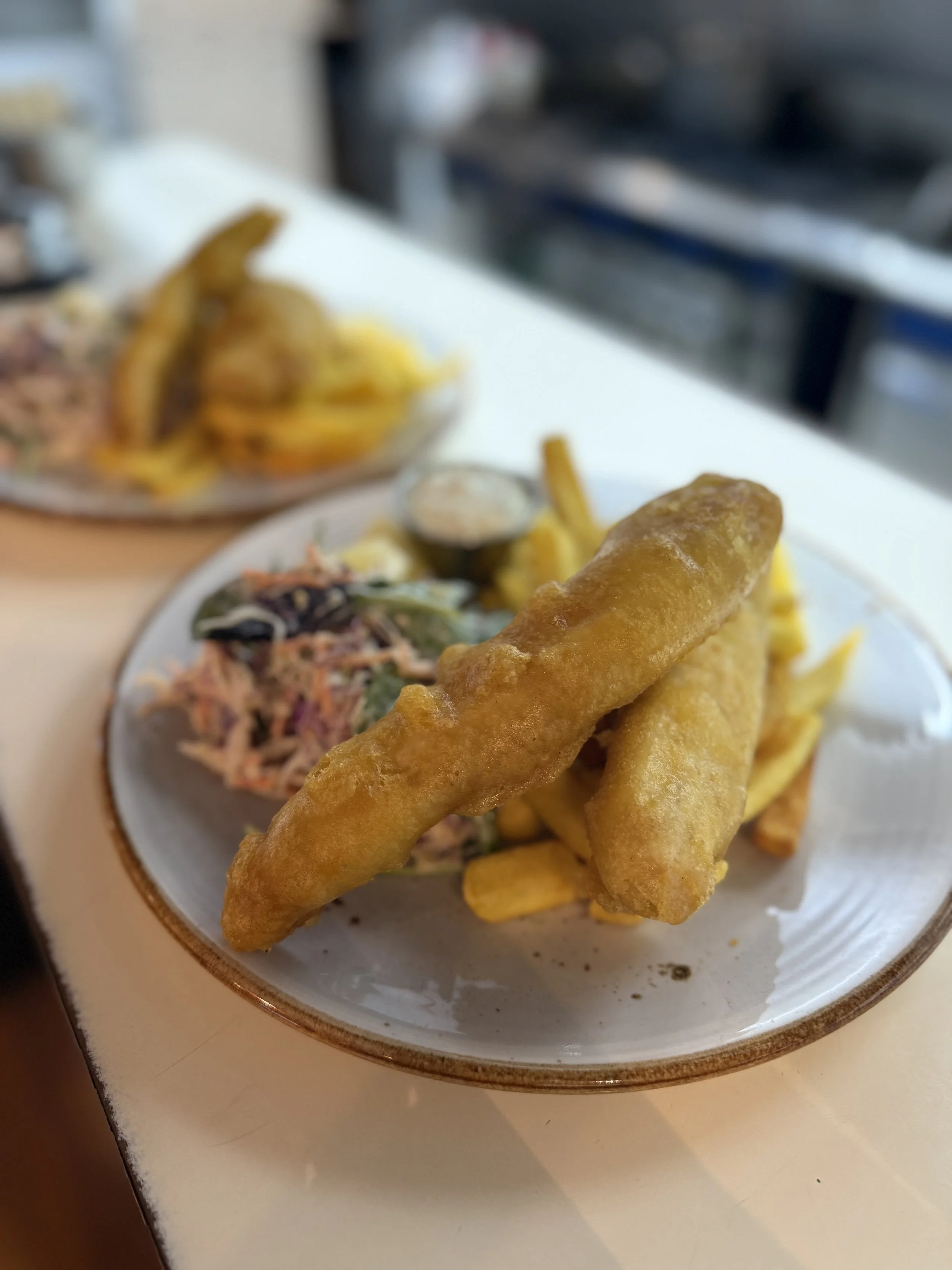 Close-up of a plate with fried fish, French fries, coleslaw, and a small container of dipping sauce, on a white table.