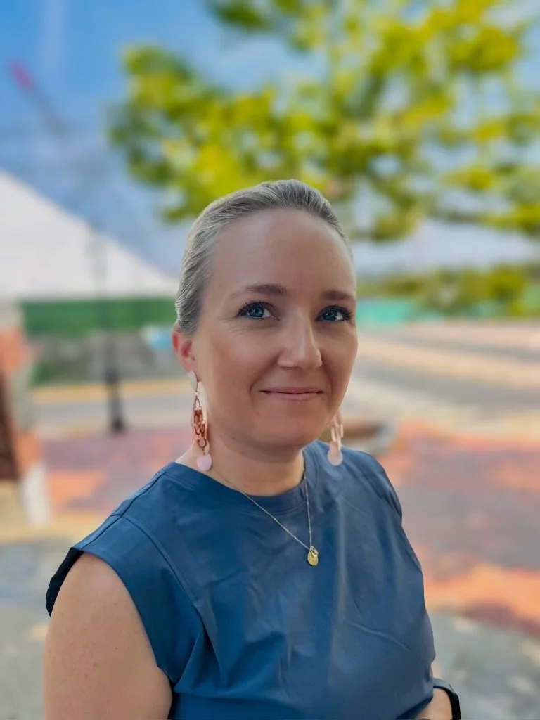 A person with blond hair is outside, wearing a blue top and earrings, smiling at the camera.