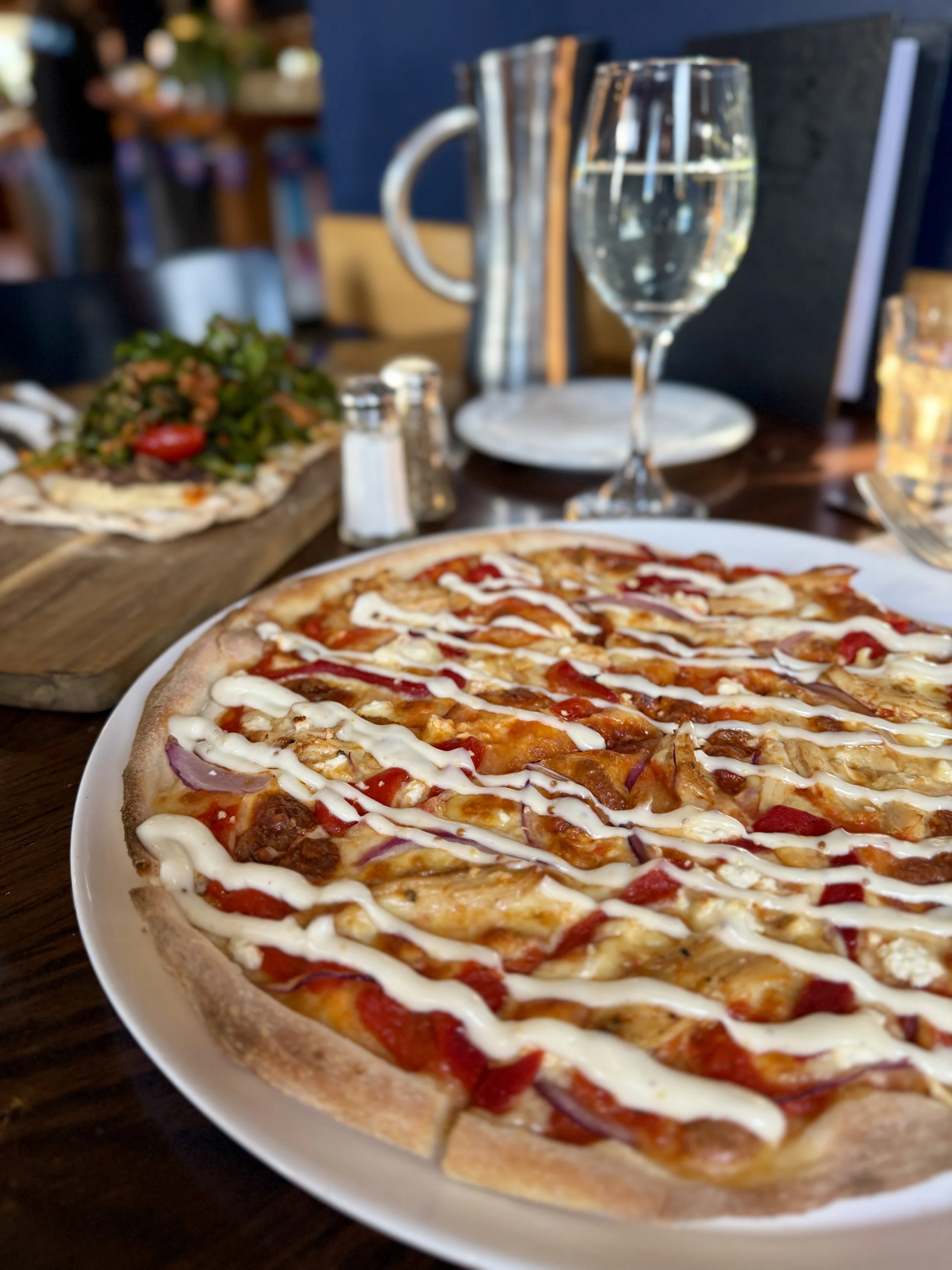 A close-up of a pizza with toppings like red onions, red peppers, and melted cheese with drizzle on top, on a white plate. In the background, there is a glass of water, a small salad, and other restaurant settings.