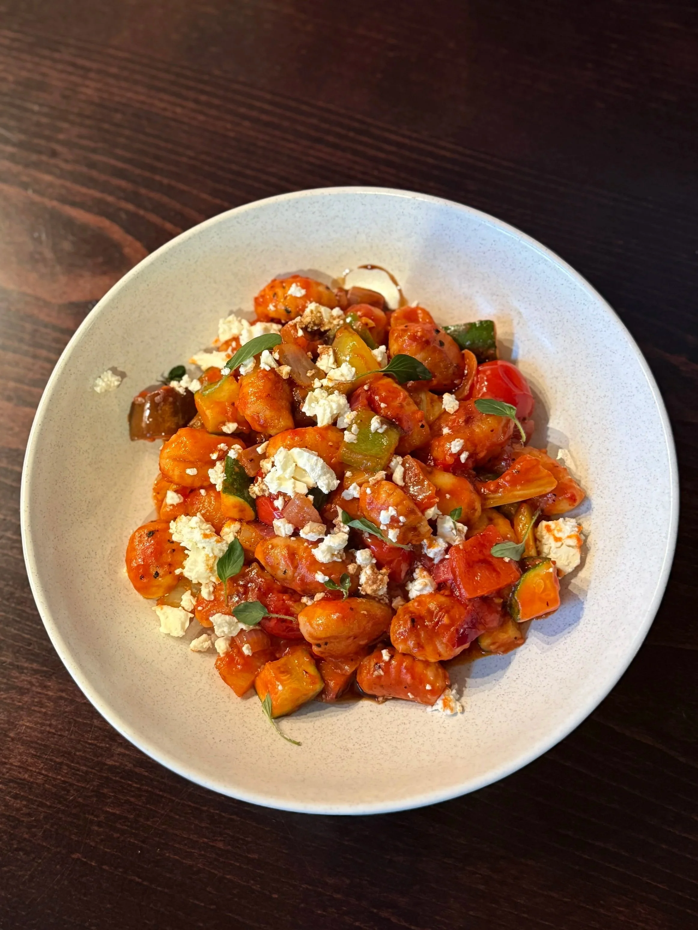 A white bowl filled with hot chicken and vegetable stew, topped with crumbled cheese and fresh herbs, on a dark wooden table.