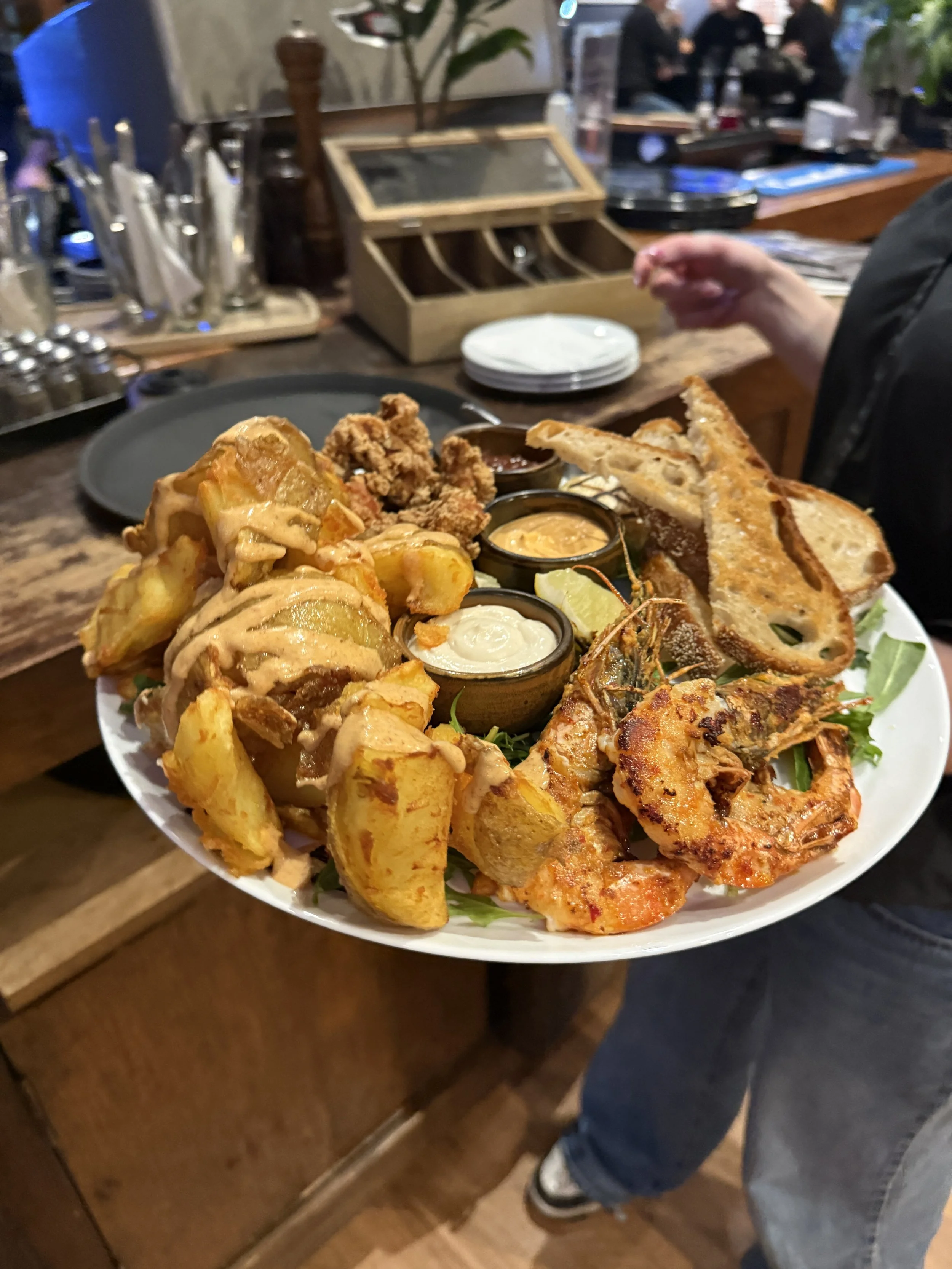 A large plate of assorted fried foods including shrimp, chicken wings, potato wedges, breaded fish, and dipping sauces in small containers, on a wooden table in a restaurant.