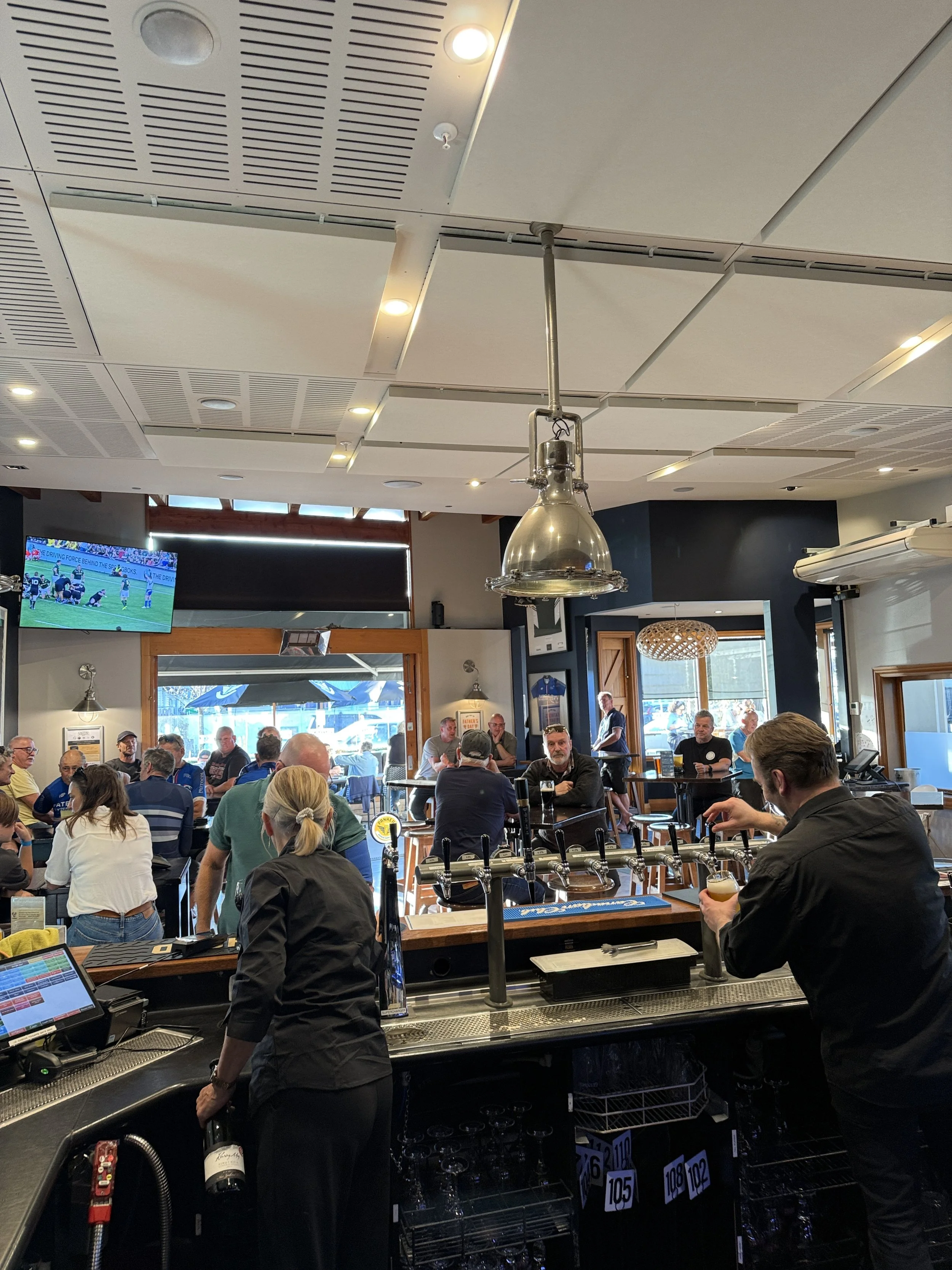 Inside a busy restaurant or bar with numerous patrons seated and standing. The image shows a counter with a bartender or server pouring drinks, a TV screen showing a sports game, and various bar equipment. The ceiling has modern lighting fixtures, and the environment appears lively and social.