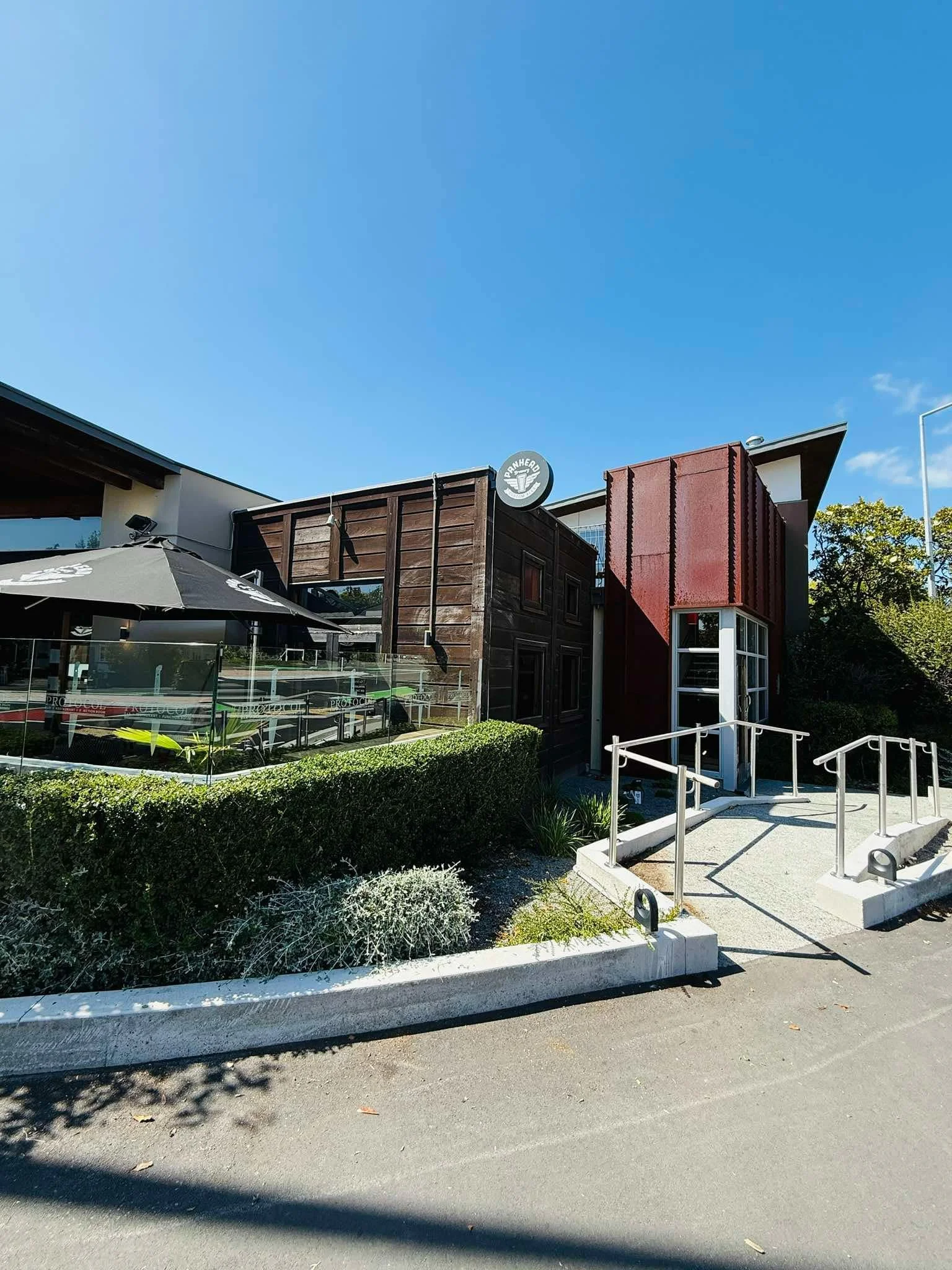 Exterior of a modern building with wooden and metal accents, a sign that reads 'PRAHED', outdoor seating area with a large umbrella, greenery, and a sidewalk with stairs and a railing under a clear blue sky.