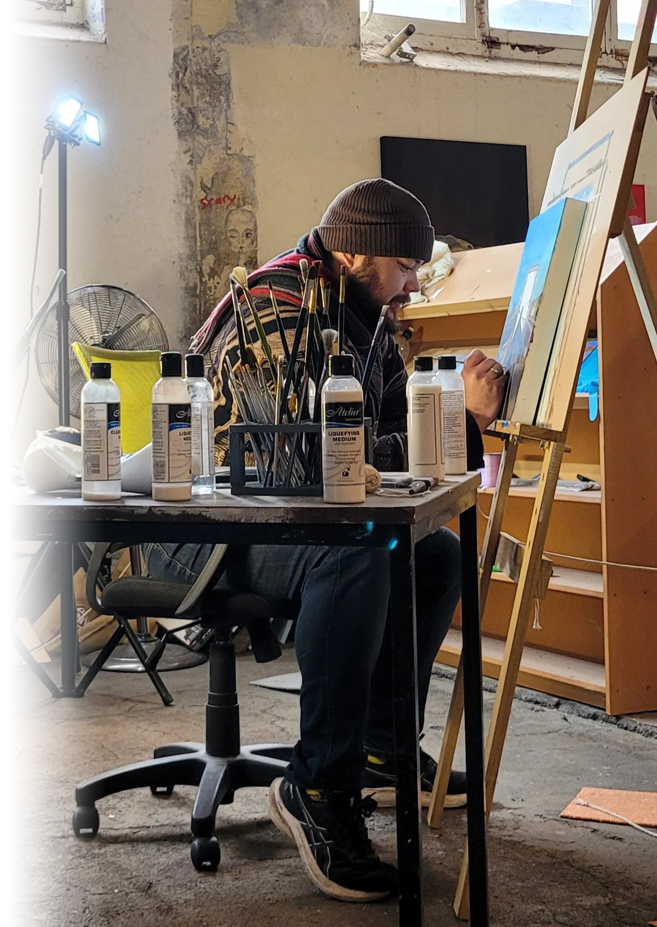 The artist, Rowan Love, painting at an easel in a studio surrounded by paintbrushes, acrylic mediums, and work lights