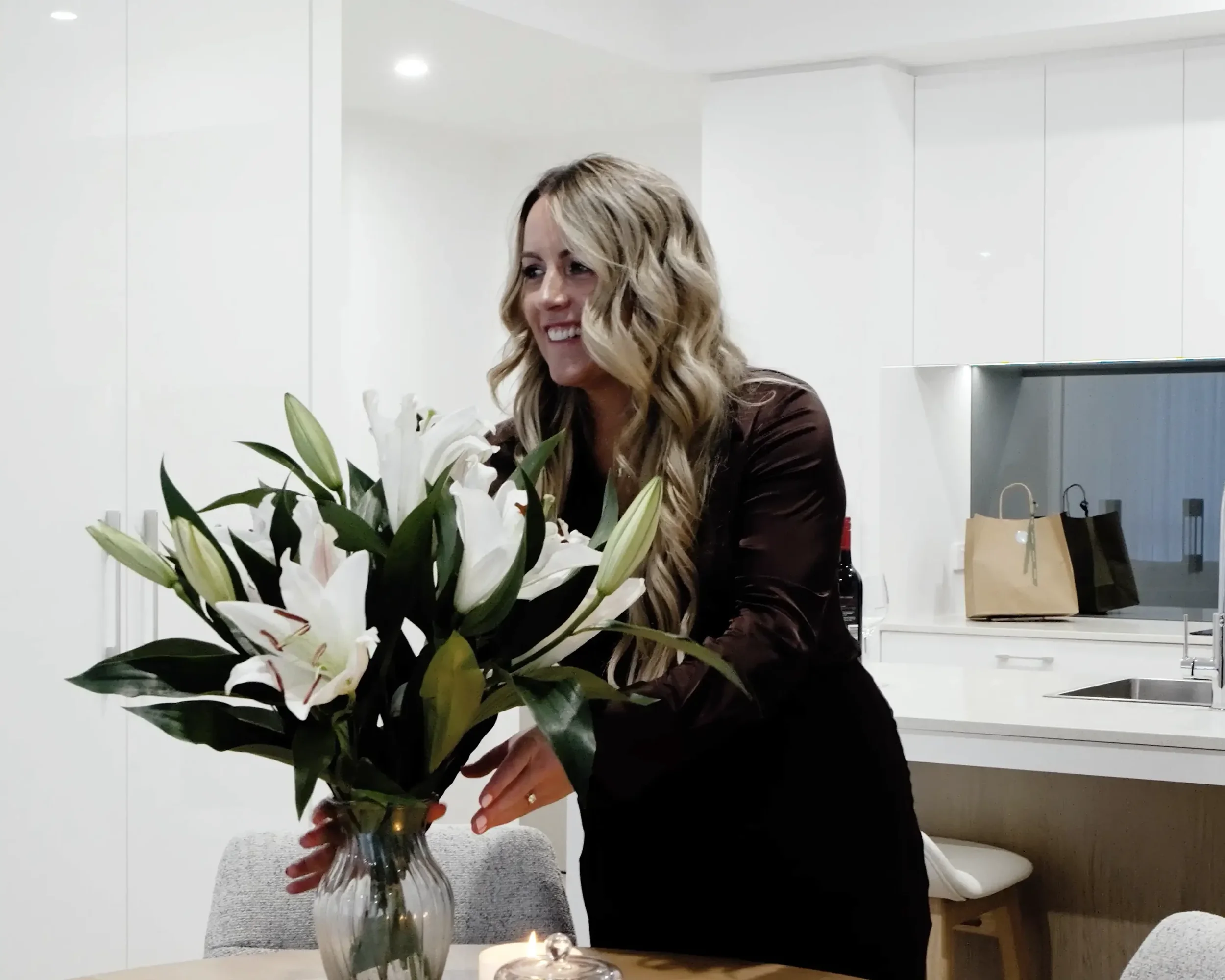 A woman with long wavy blonde hair smiling while arranging white lilies in a glass vase on a kitchen table.